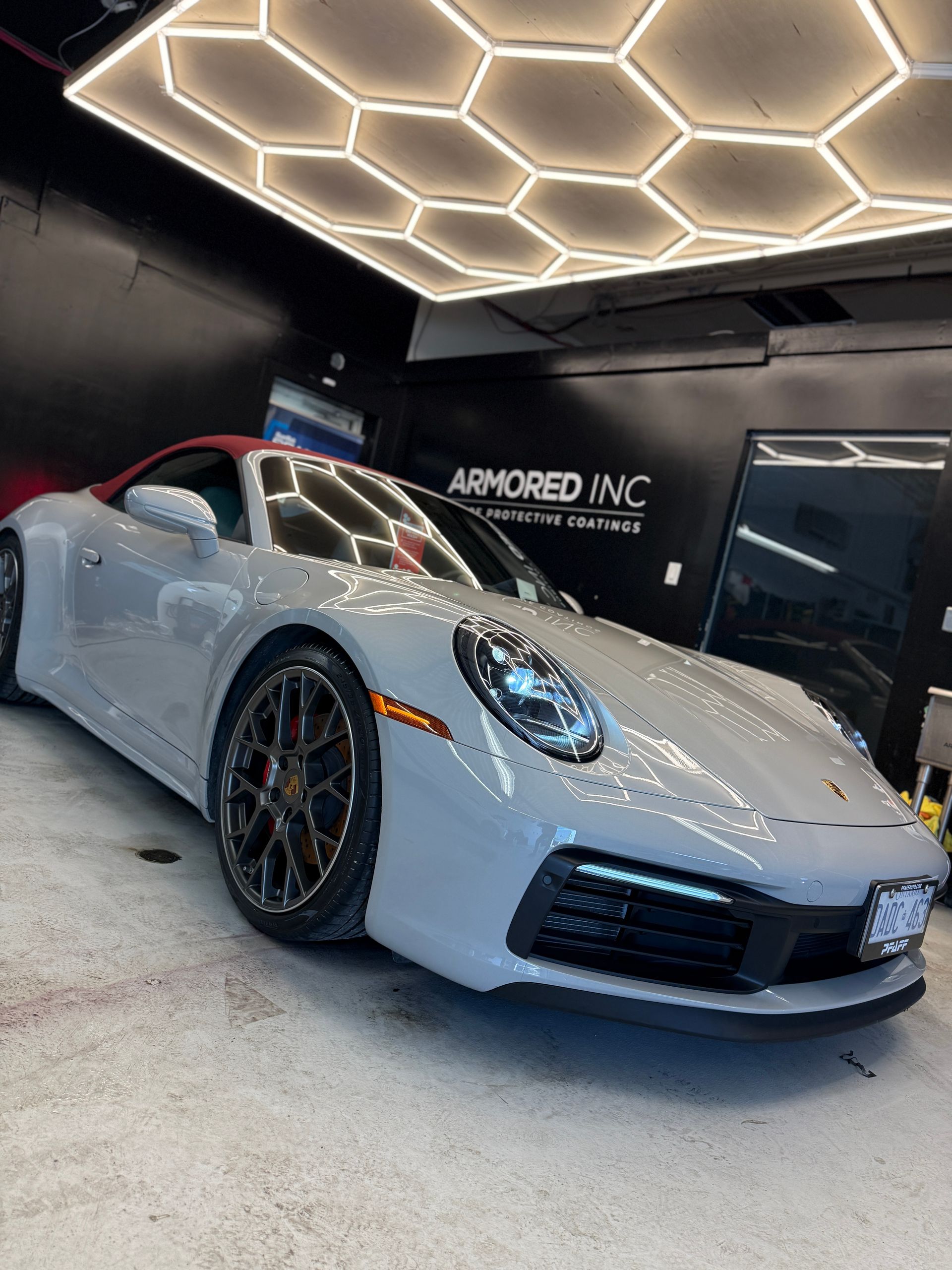 A light gray Porsche convertible with a red top parked in an indoor garage with geometric hexagon ceiling lights.