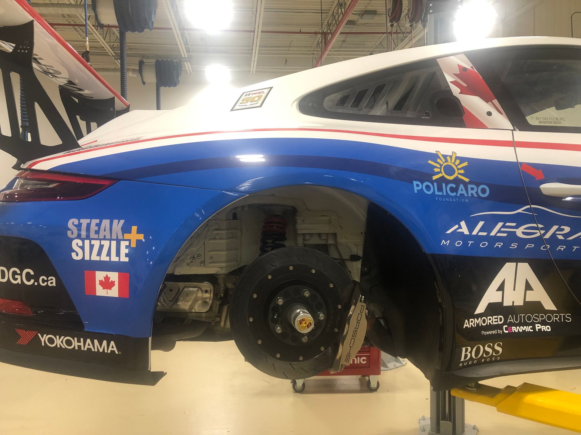 A race car with blue and white livery, Canadian flags, and sponsors sits in a workshop without its rear tire.