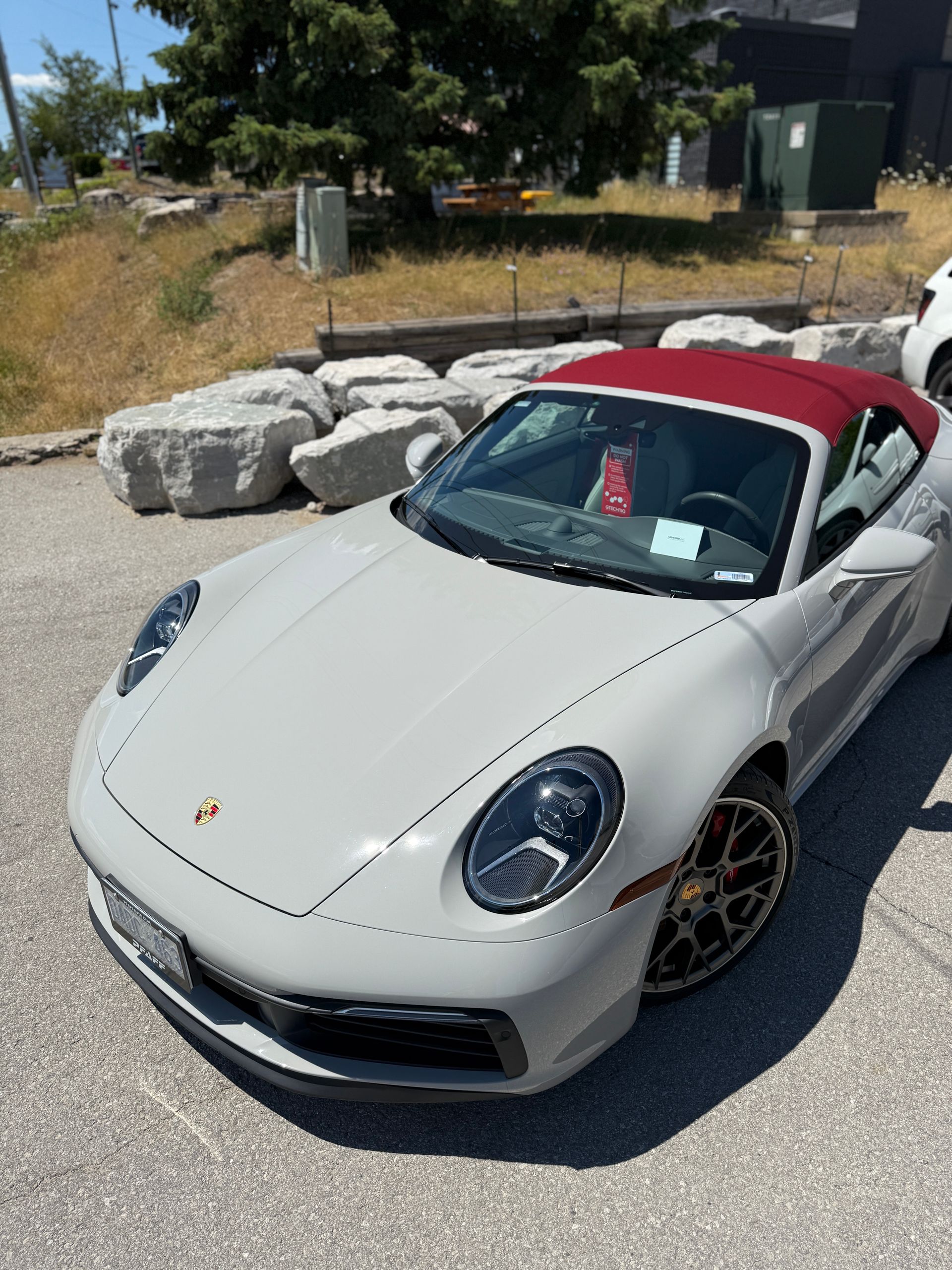 A grey Porsche convertible with a red soft top parked on a gravel lot near a tree and large landscaping rocks.