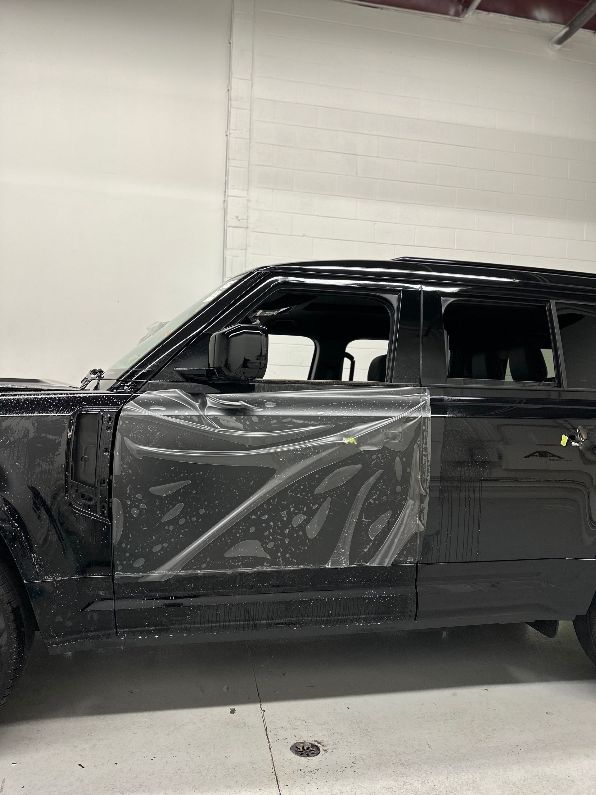 A black car parked indoors with a clear protective film partially applied to the driver’s side door.