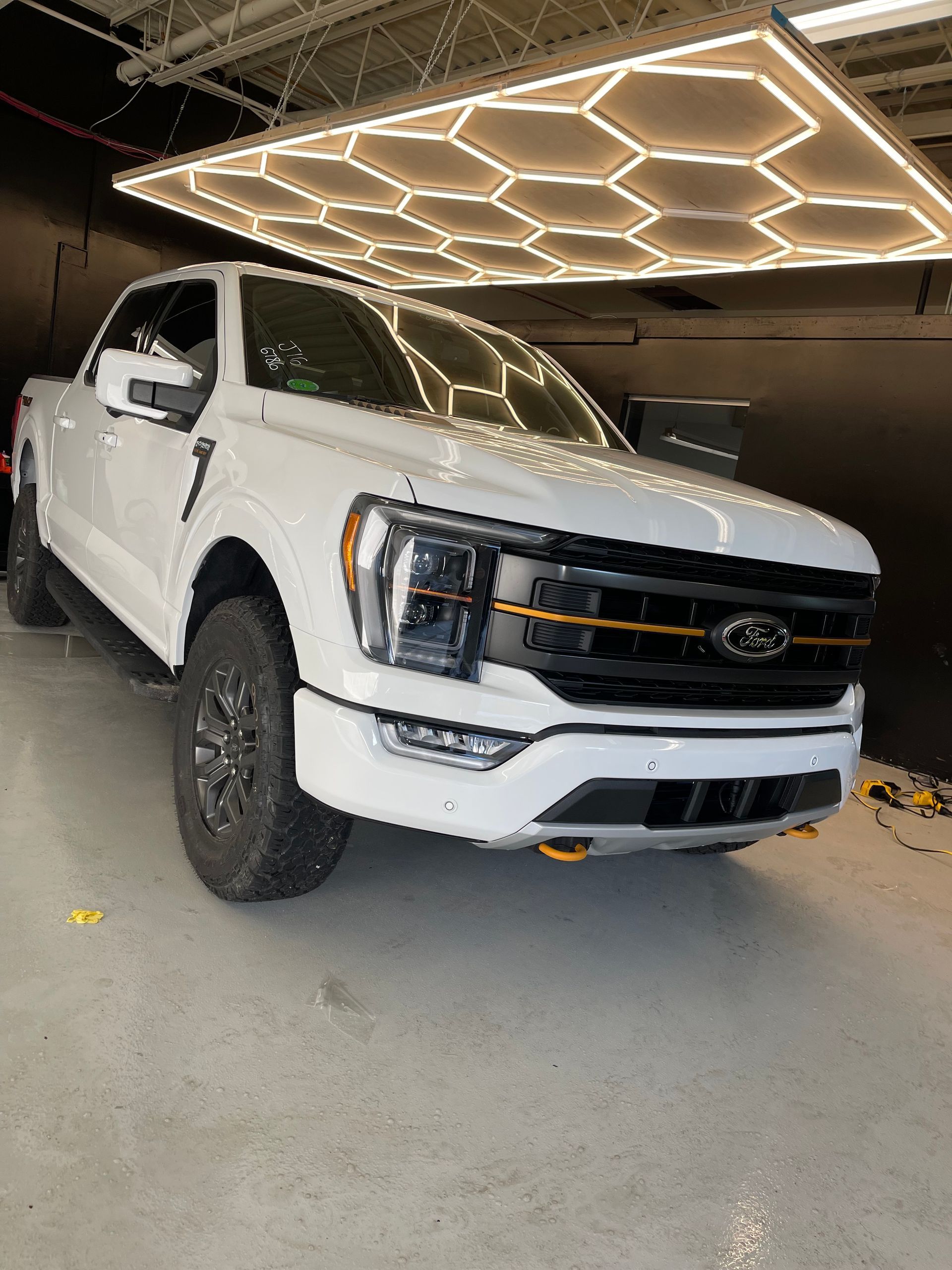 A white Ford F-150 pickup truck with black trim and orange accents parked in a garage with hexagonal ceiling lights.
