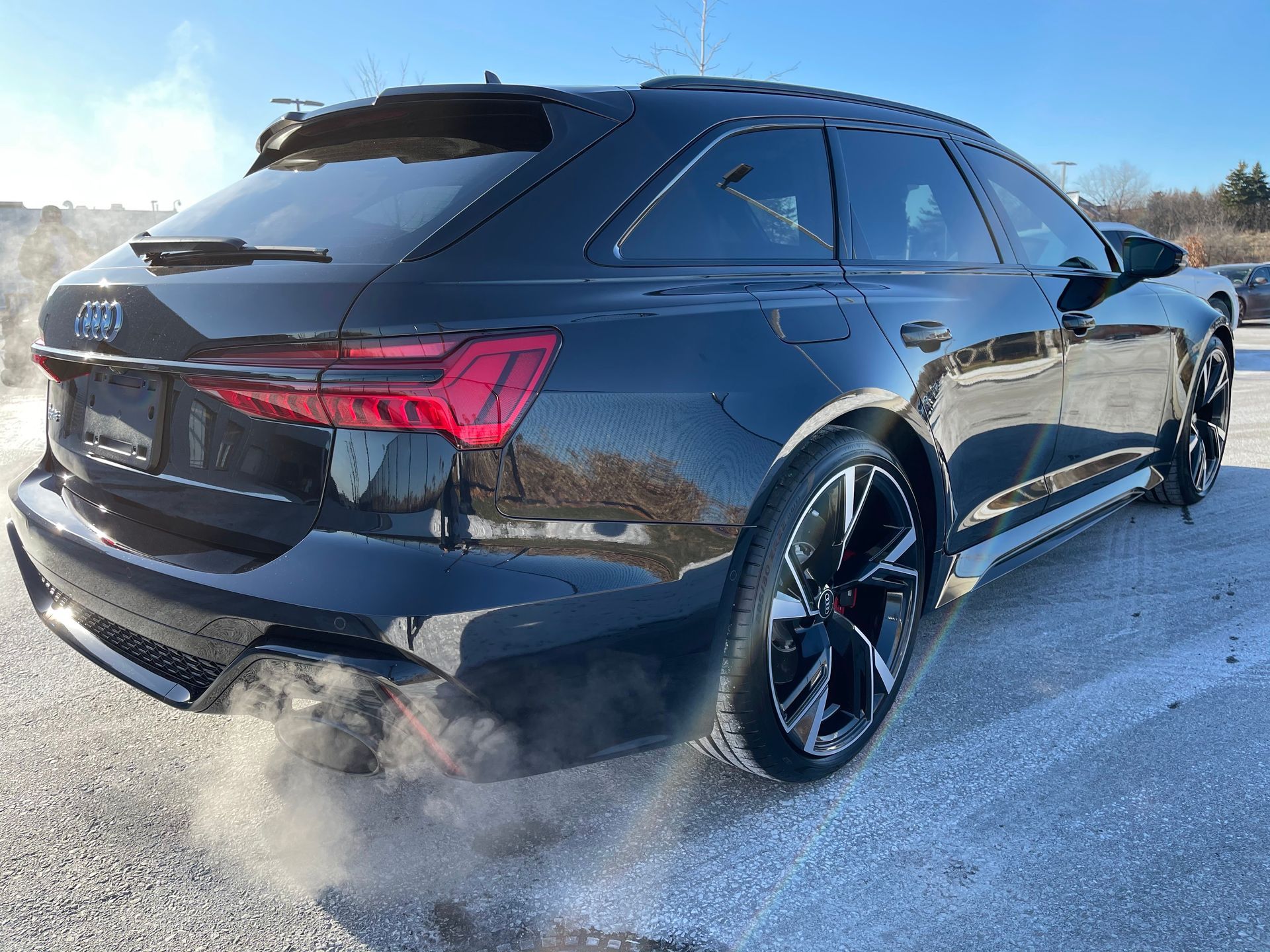 A black Audi station wagon parked on a snowy lot, emitting exhaust fumes from the rear.