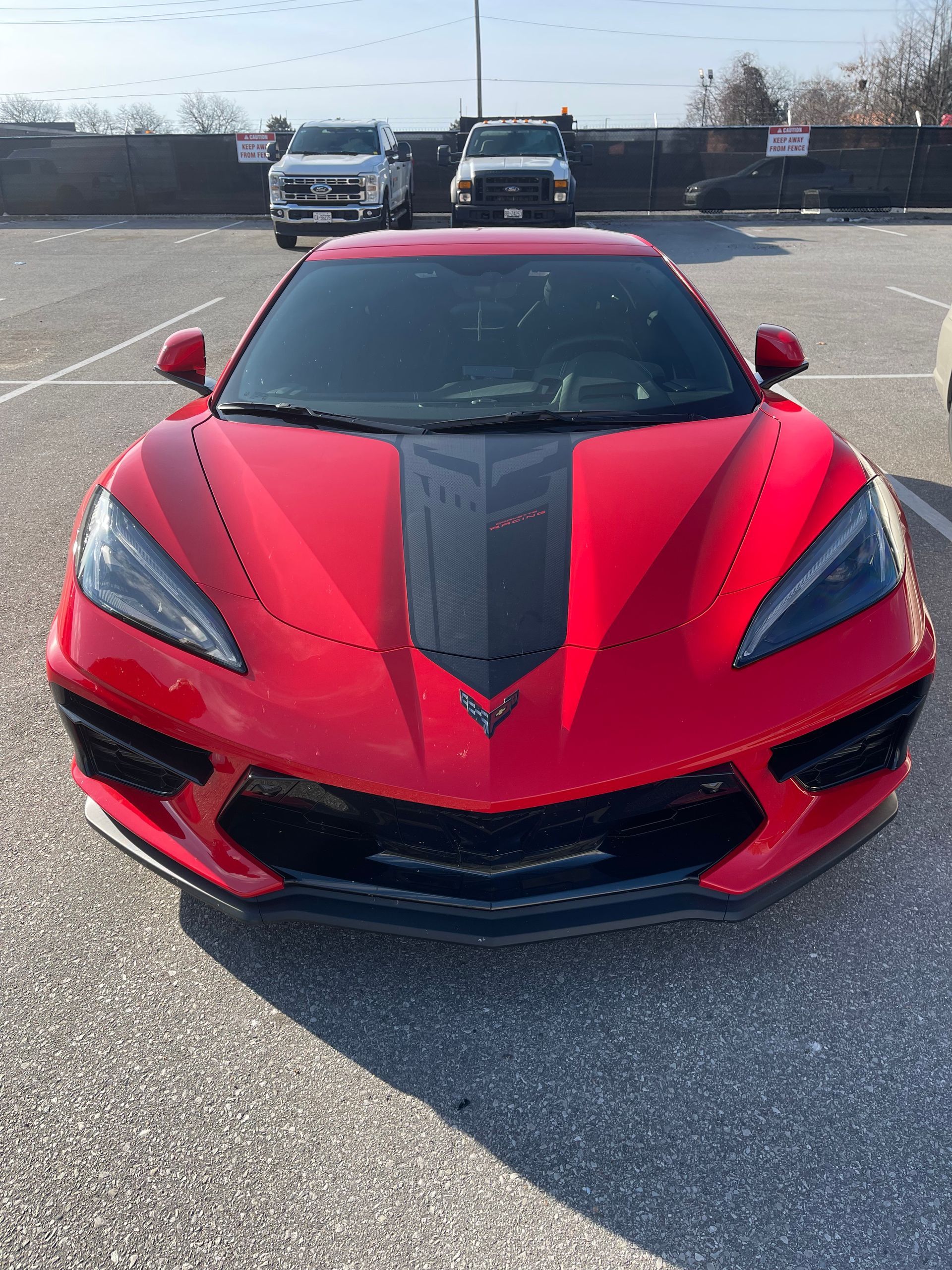 A red Chevrolet Corvette parked in a sunny outdoor lot, showing its front view and a central black racing stripe.