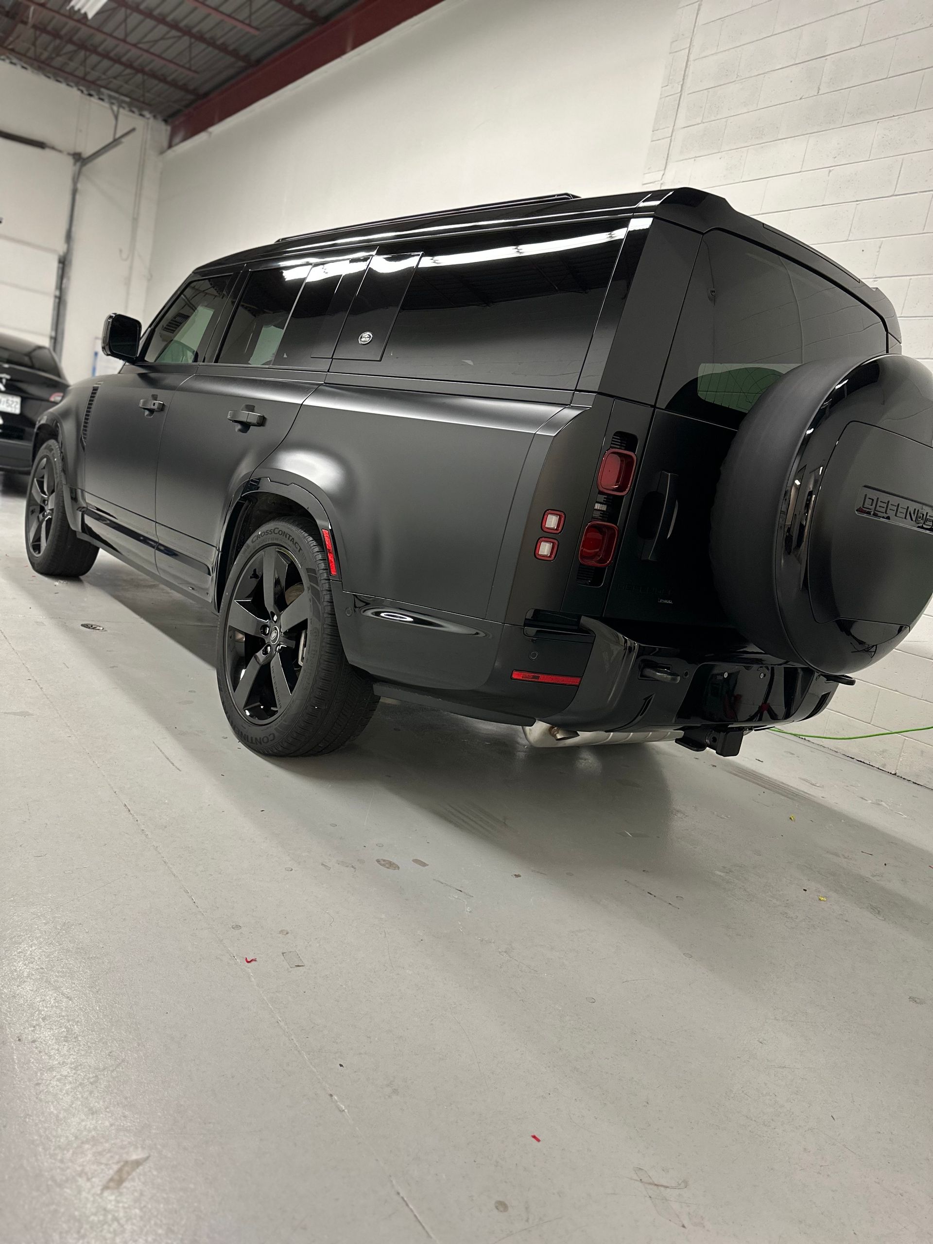 A matte black Land Rover Defender in an indoor garage, seen from a rear-three-quarter angle with a rear-mounted spare tire.