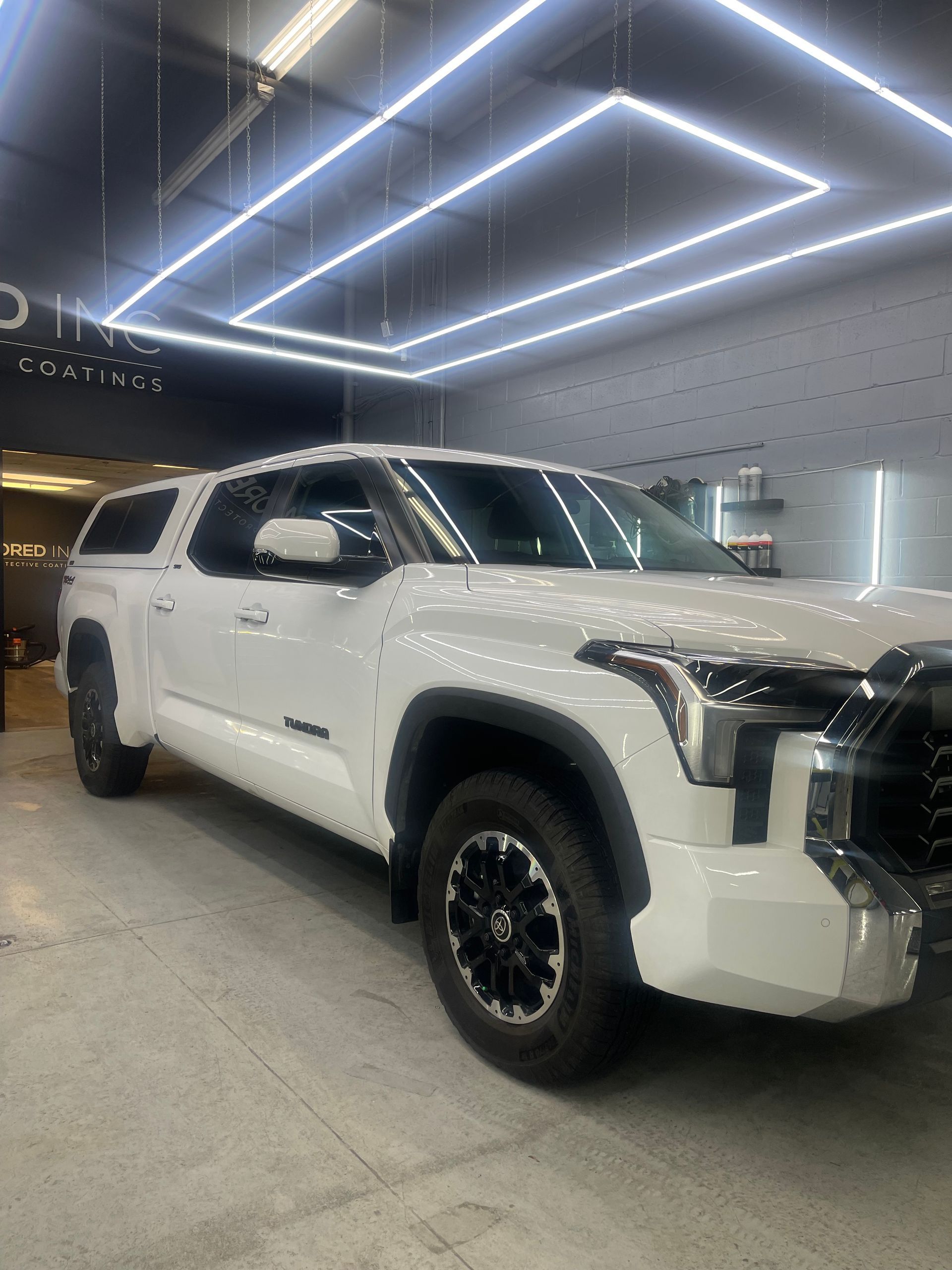 A white Toyota Tundra truck with a camper shell parked in a well-lit showroom with overhead geometric LED lights.