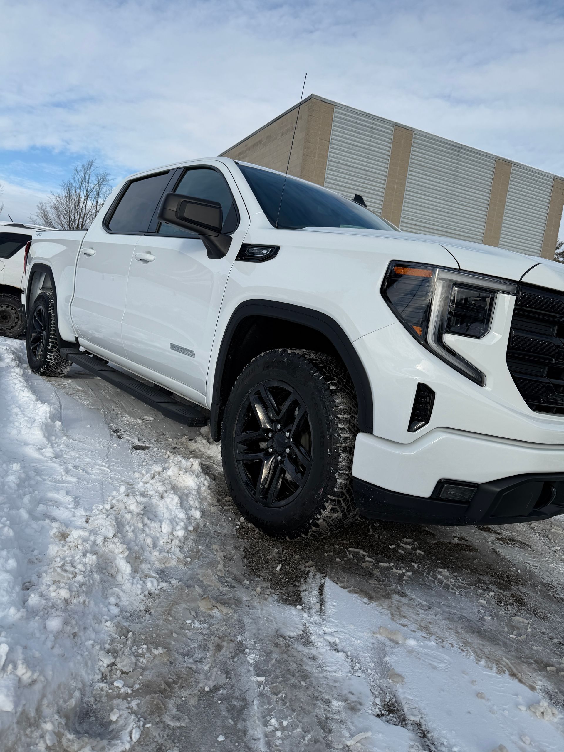 A white GMC Sierra pickup truck with black wheels parked in a snowy lot.