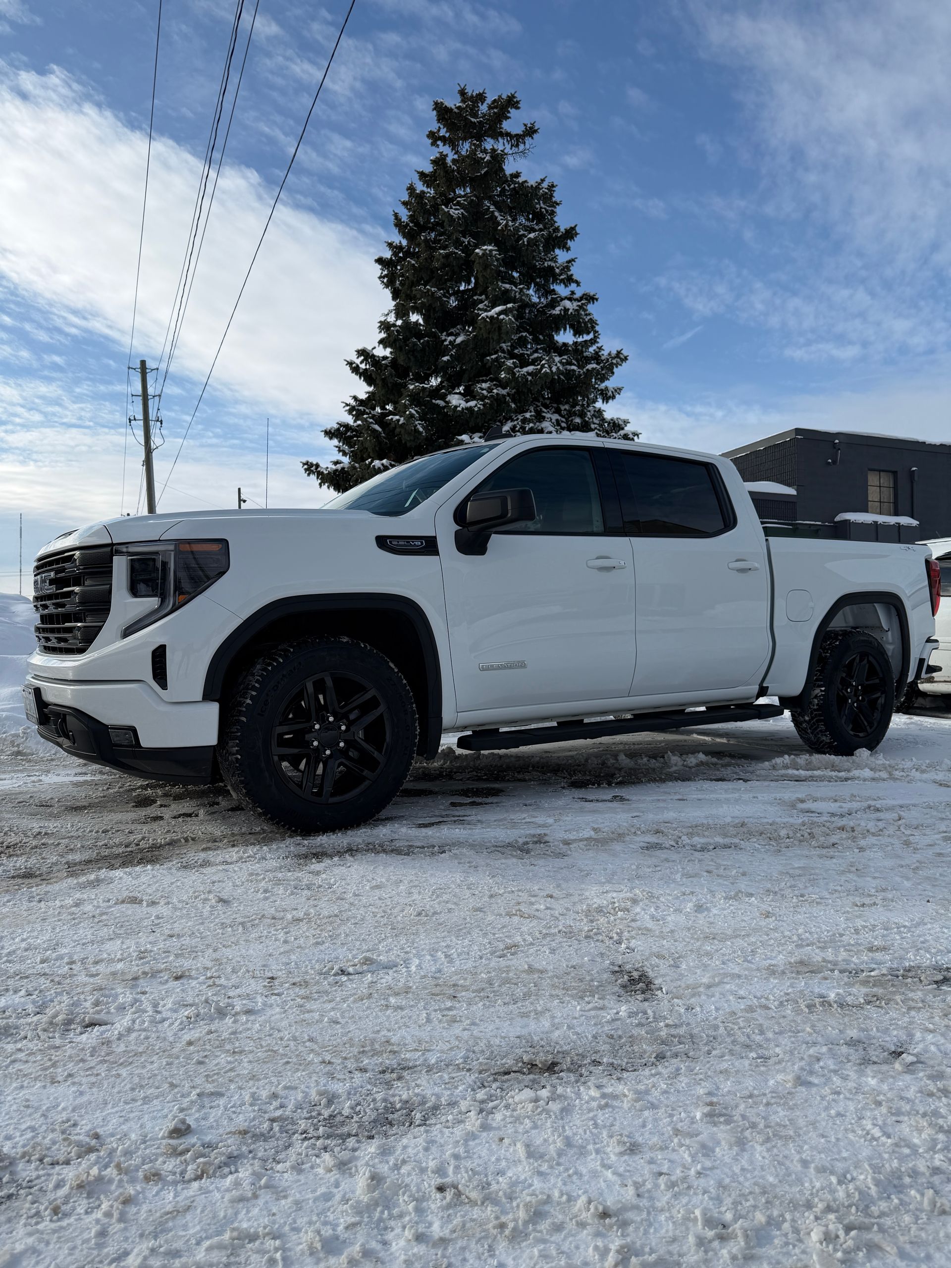 A white GMC Sierra pickup truck with black wheels parked in a snowy lot under a blue sky with a large evergreen tree.