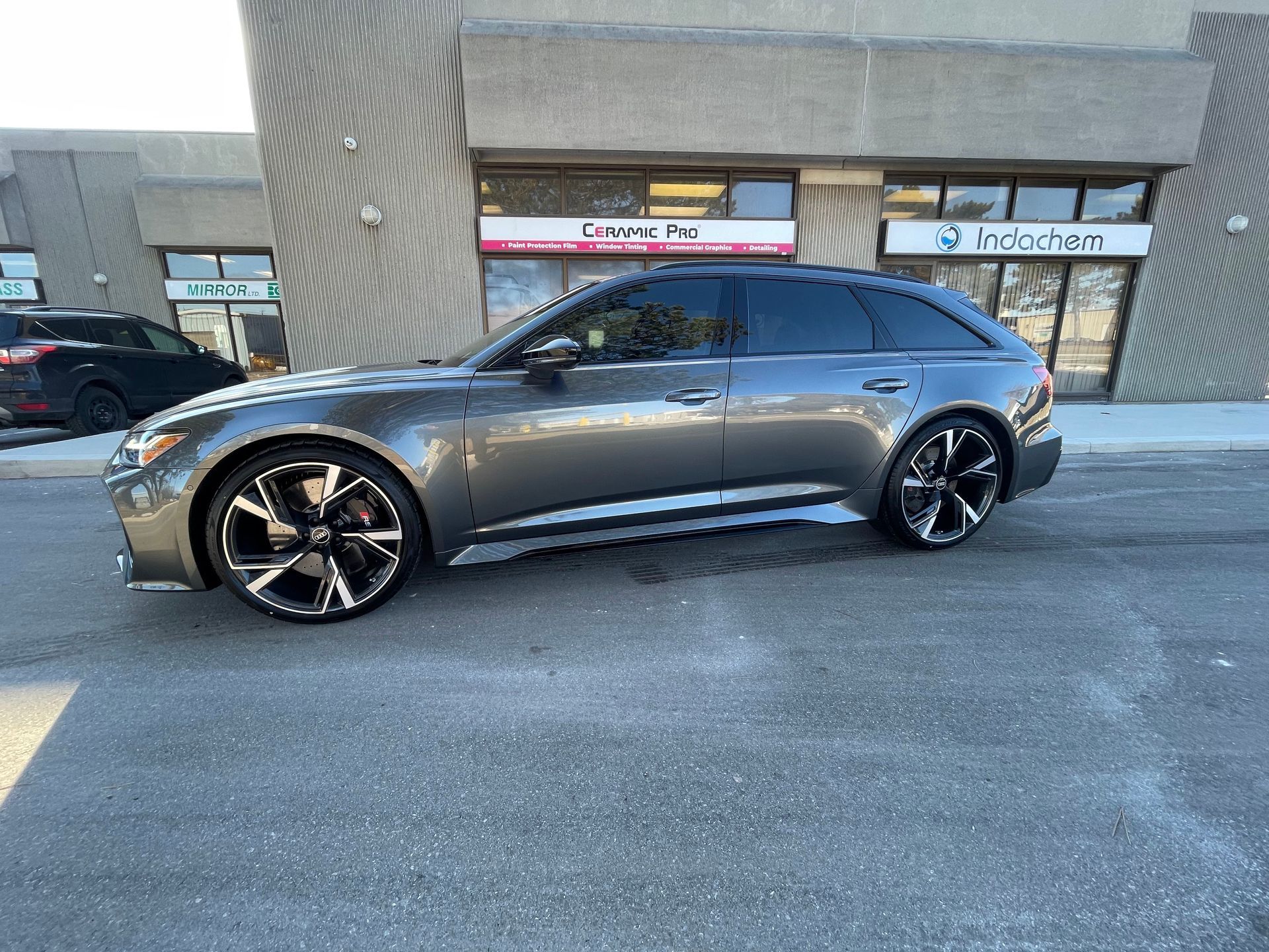 A dark gray Audi RS6 Avant wagon parked in a paved lot in front of commercial retail storefronts.