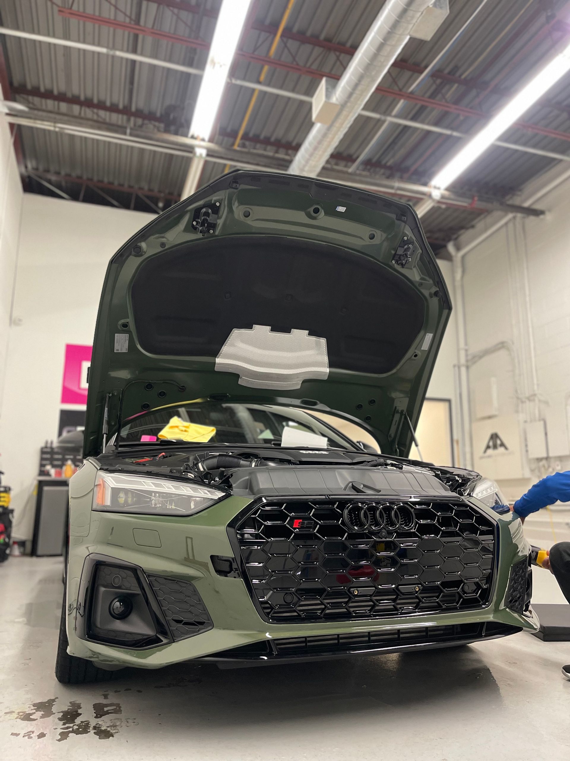An olive green Audi with its hood open, undergoing maintenance inside a brightly lit auto repair shop.