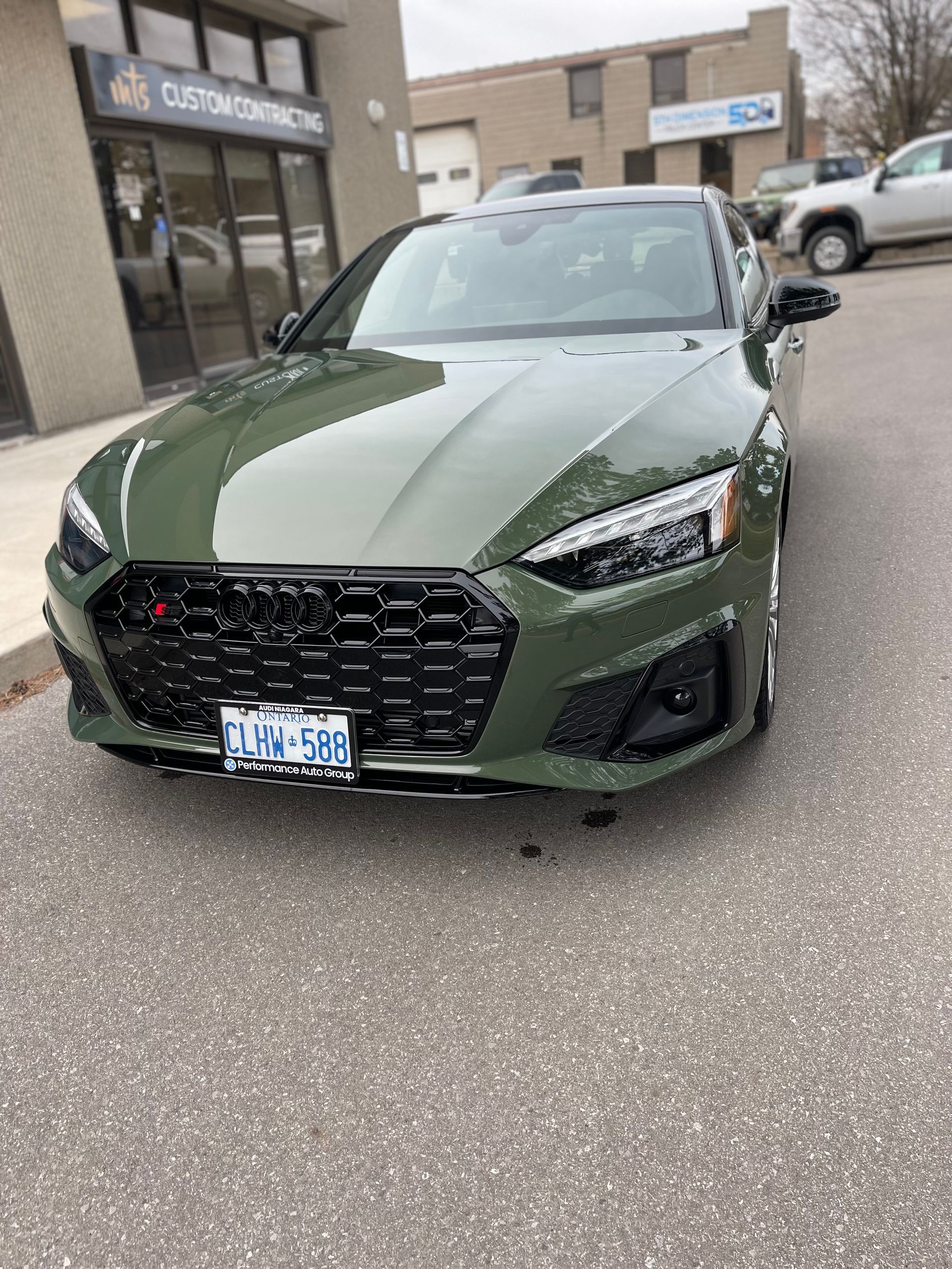 An olive green Audi S5 parked in front of a commercial building on a paved lot.