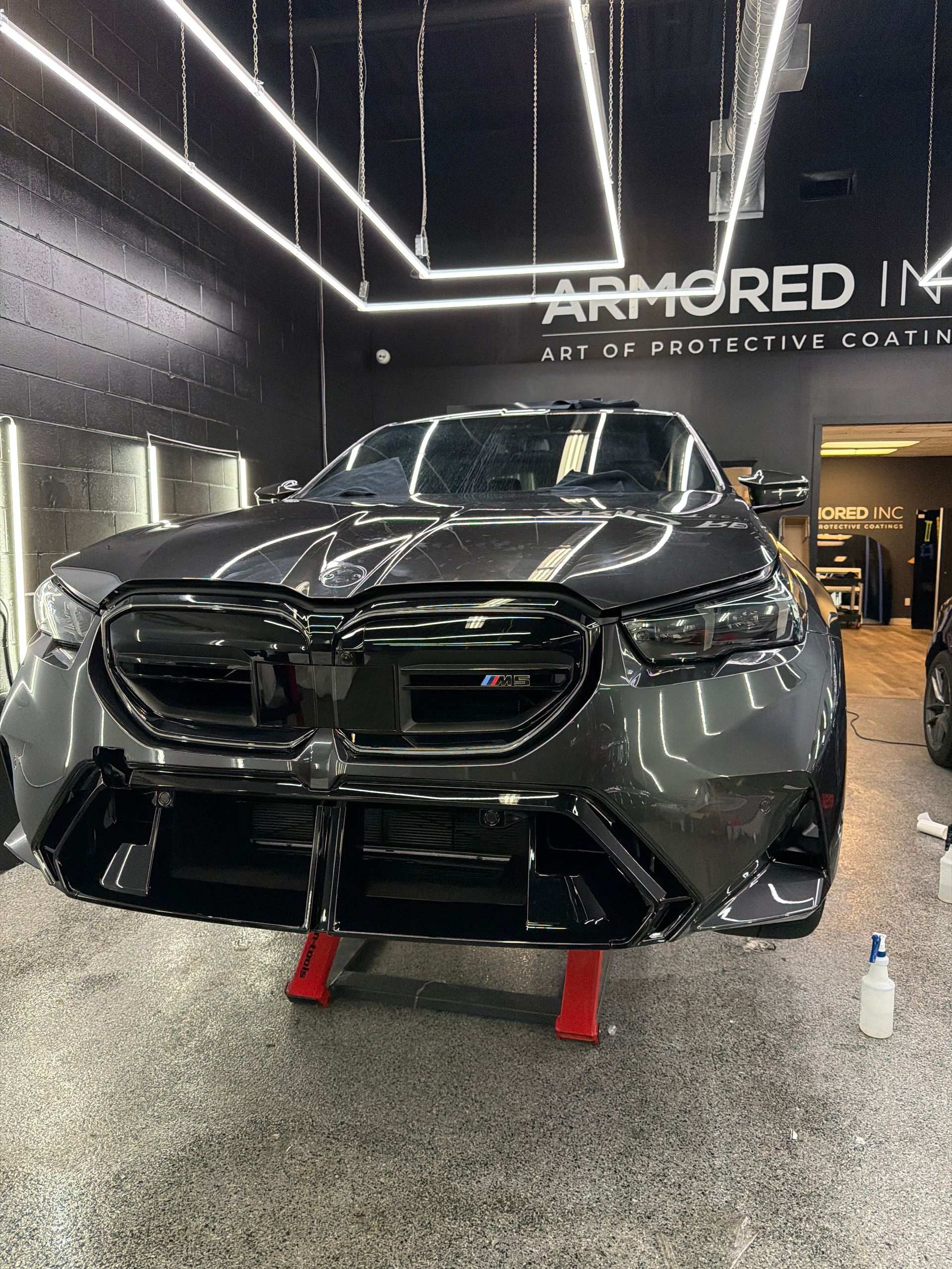 A dark gray luxury car in a professional auto detailing shop, positioned on red stands under bright overhead lighting.