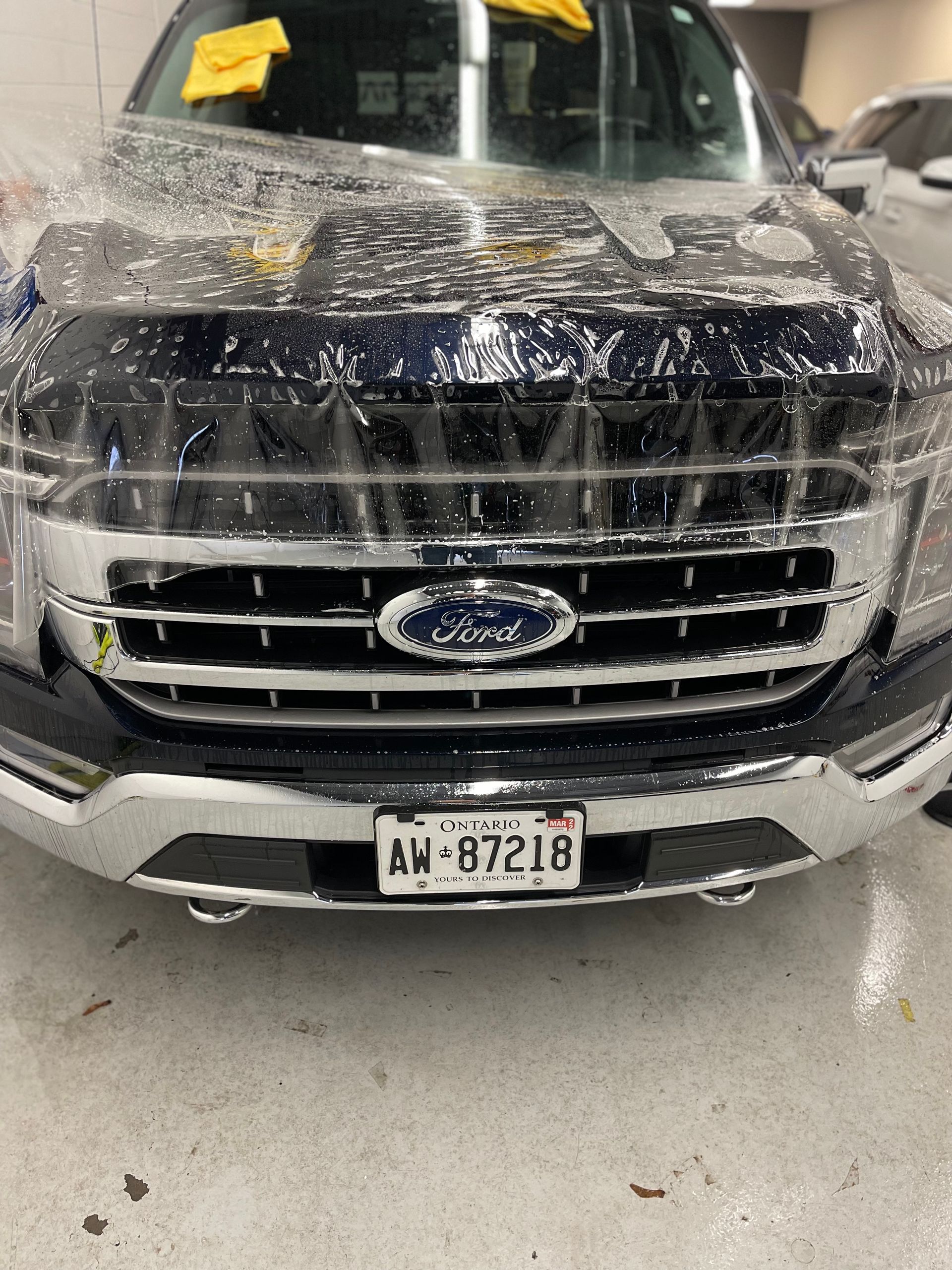 A dark blue Ford truck in an indoor service bay, partially covered in a clear protective plastic film.