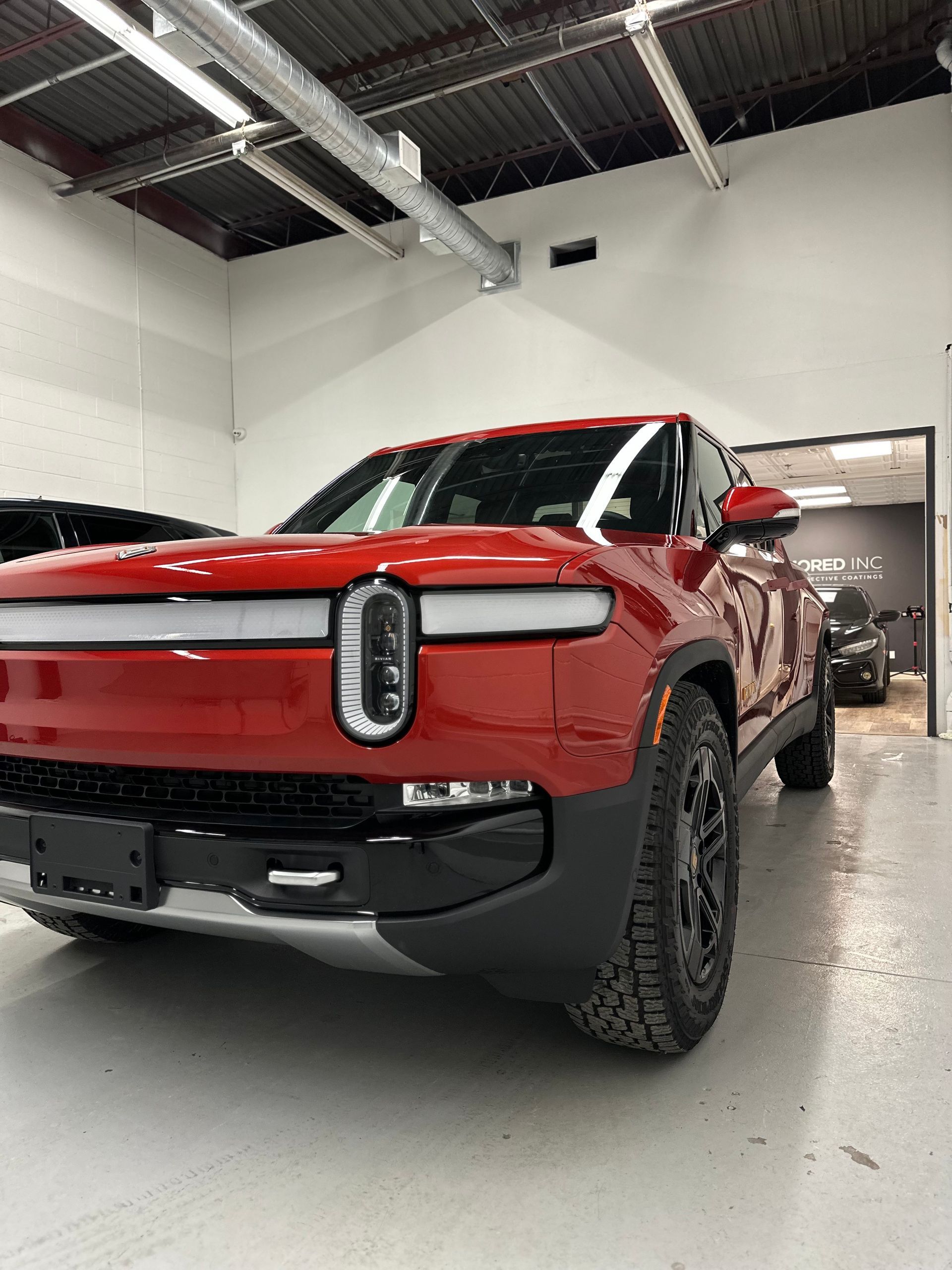 A red Rivian R1T electric pickup truck parked inside a bright, clean garage with exposed industrial ceiling pipes.