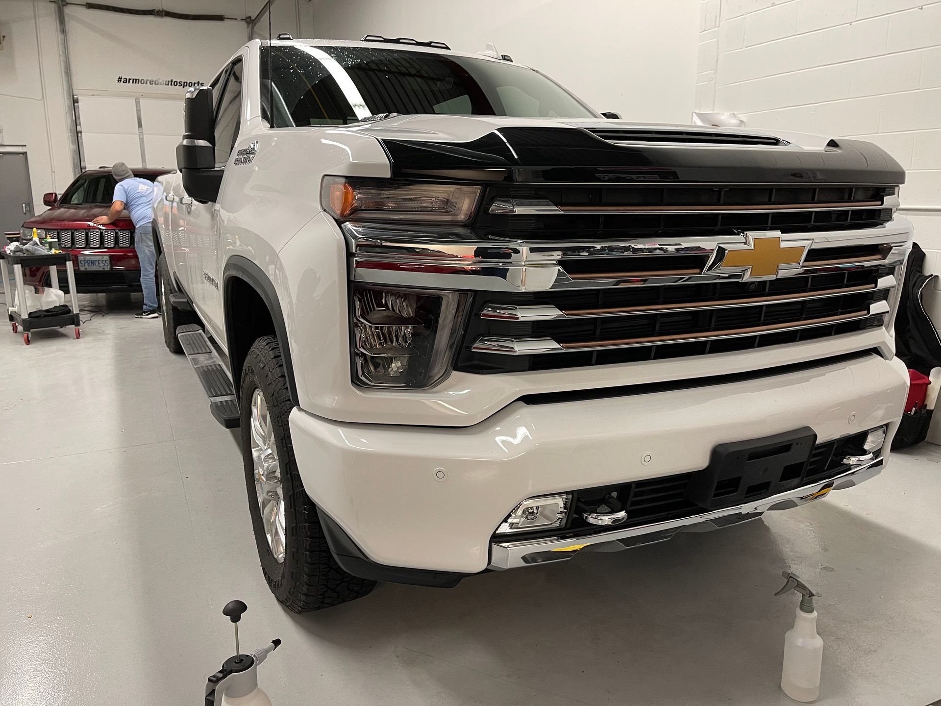 A white Chevrolet Silverado pickup truck parked in a bright garage with a mechanic working on a car in the background.