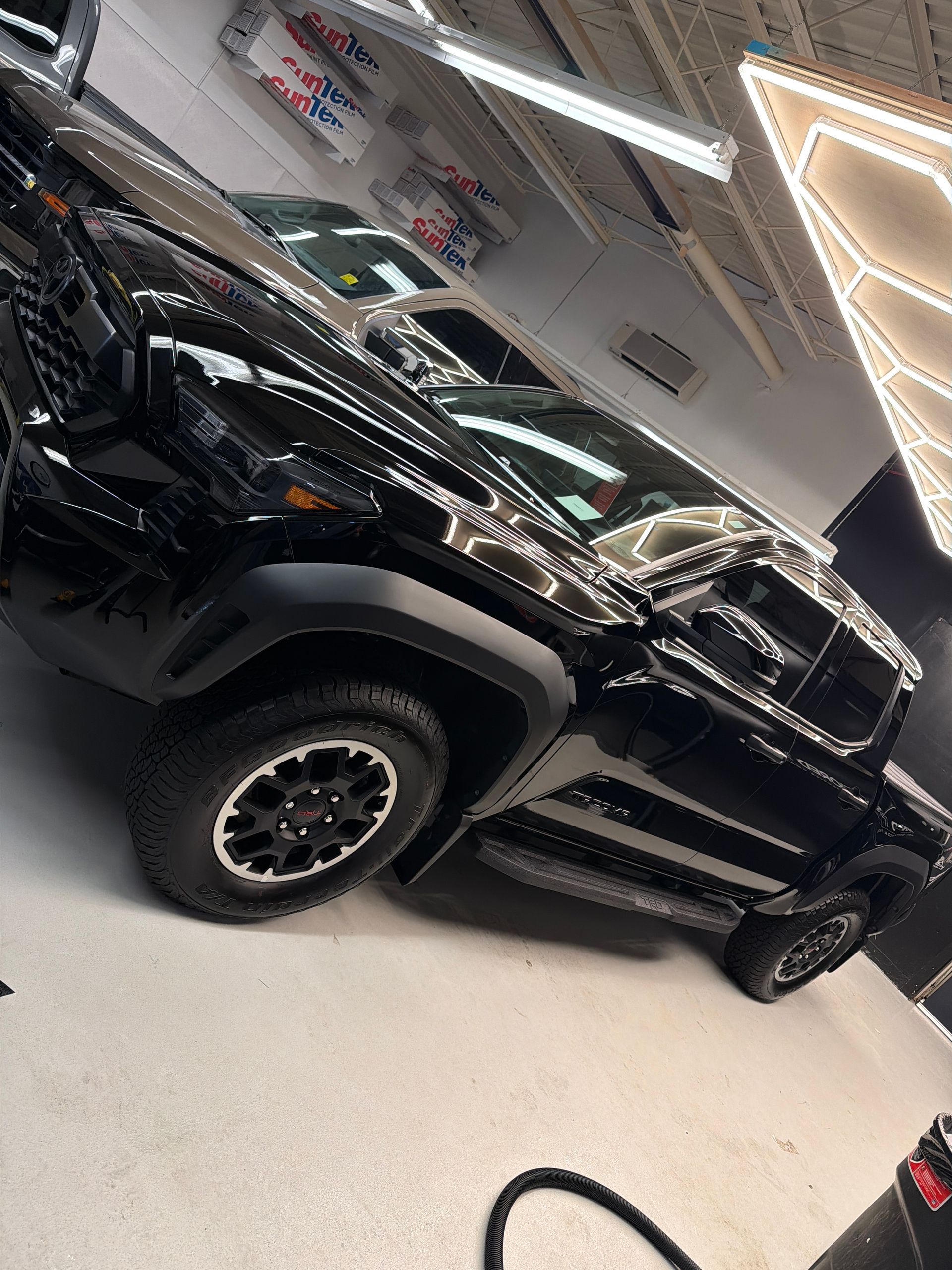 A black Toyota Tacoma pickup truck with rugged off-road tires and black wheels parked inside a brightly lit garage.