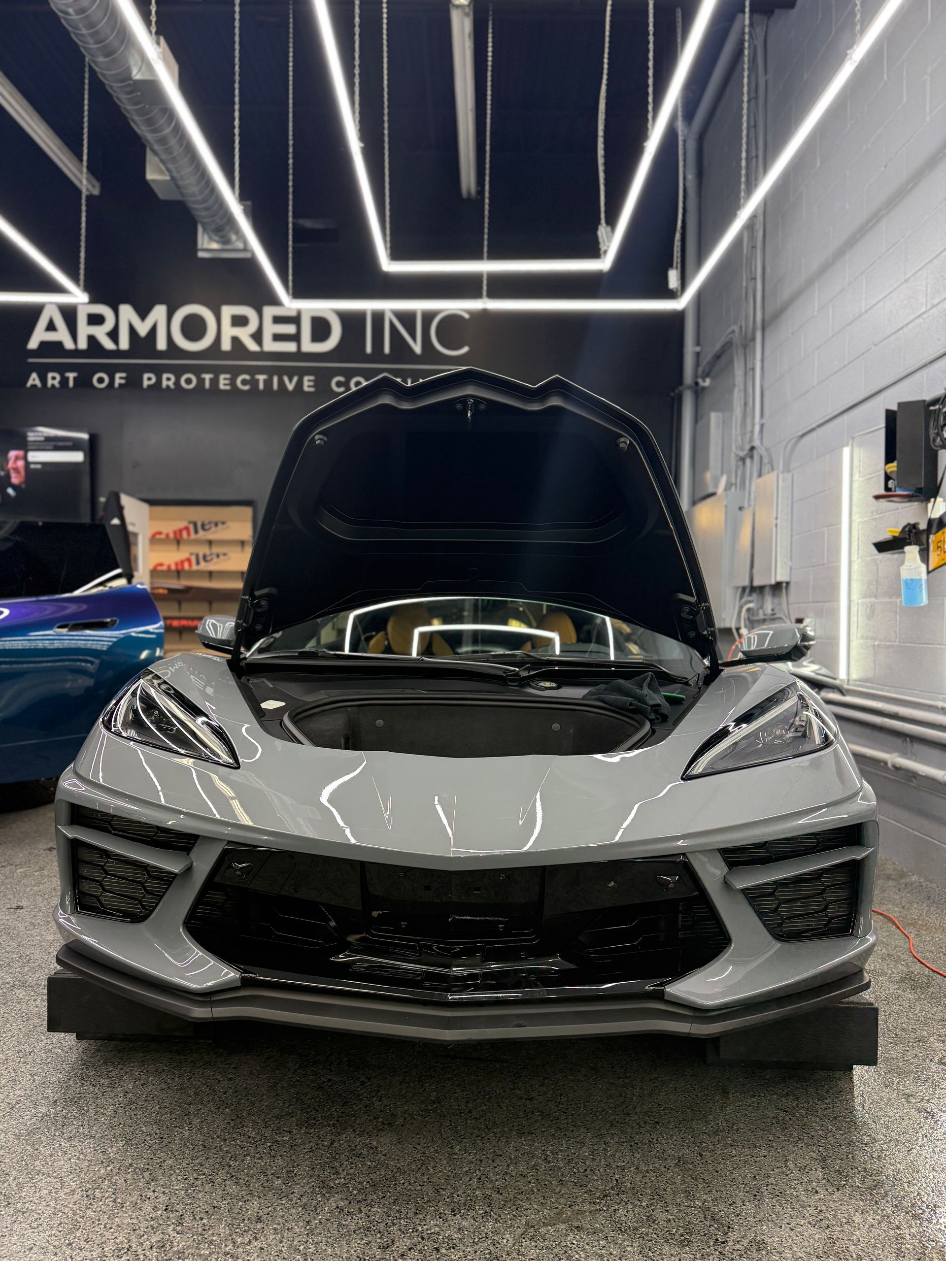 A grey Corvette with its front hood open inside an Armored Inc. shop with bright overhead lighting.