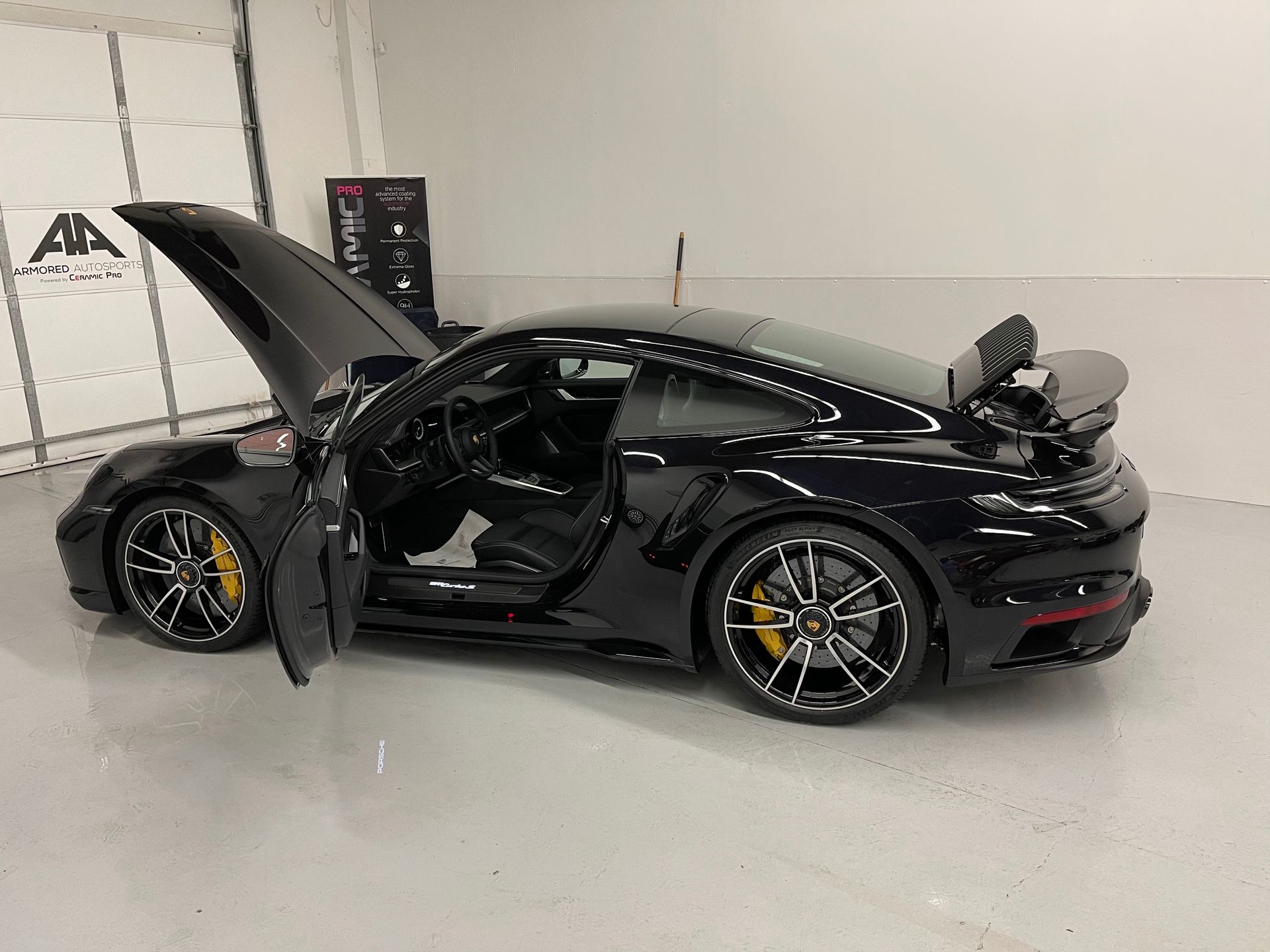A black Porsche 911 Turbo parked in a garage with its hood and rear spoiler open and driver side door ajar.