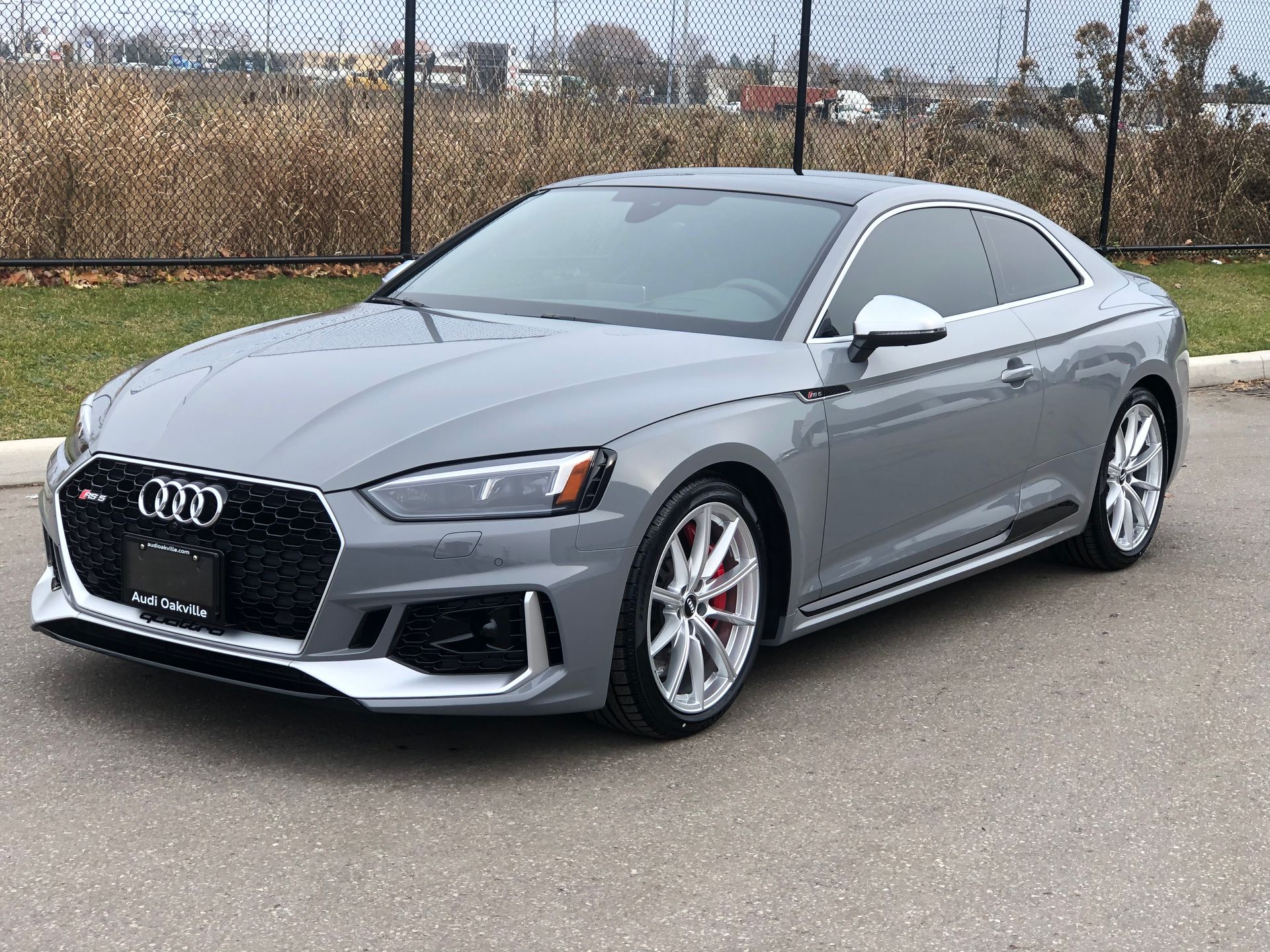 A gray Audi RS5 coupe parked on asphalt with a chain-link fence in the background.
