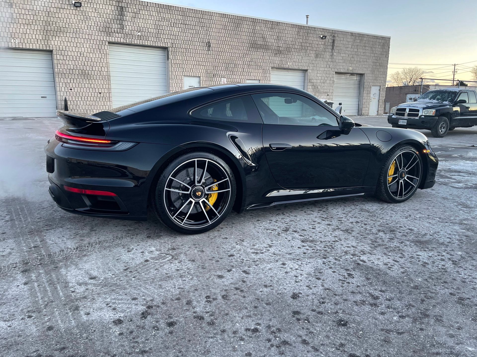 A black Porsche 911 sports car parked on a gravel lot in front of industrial buildings at sunset.