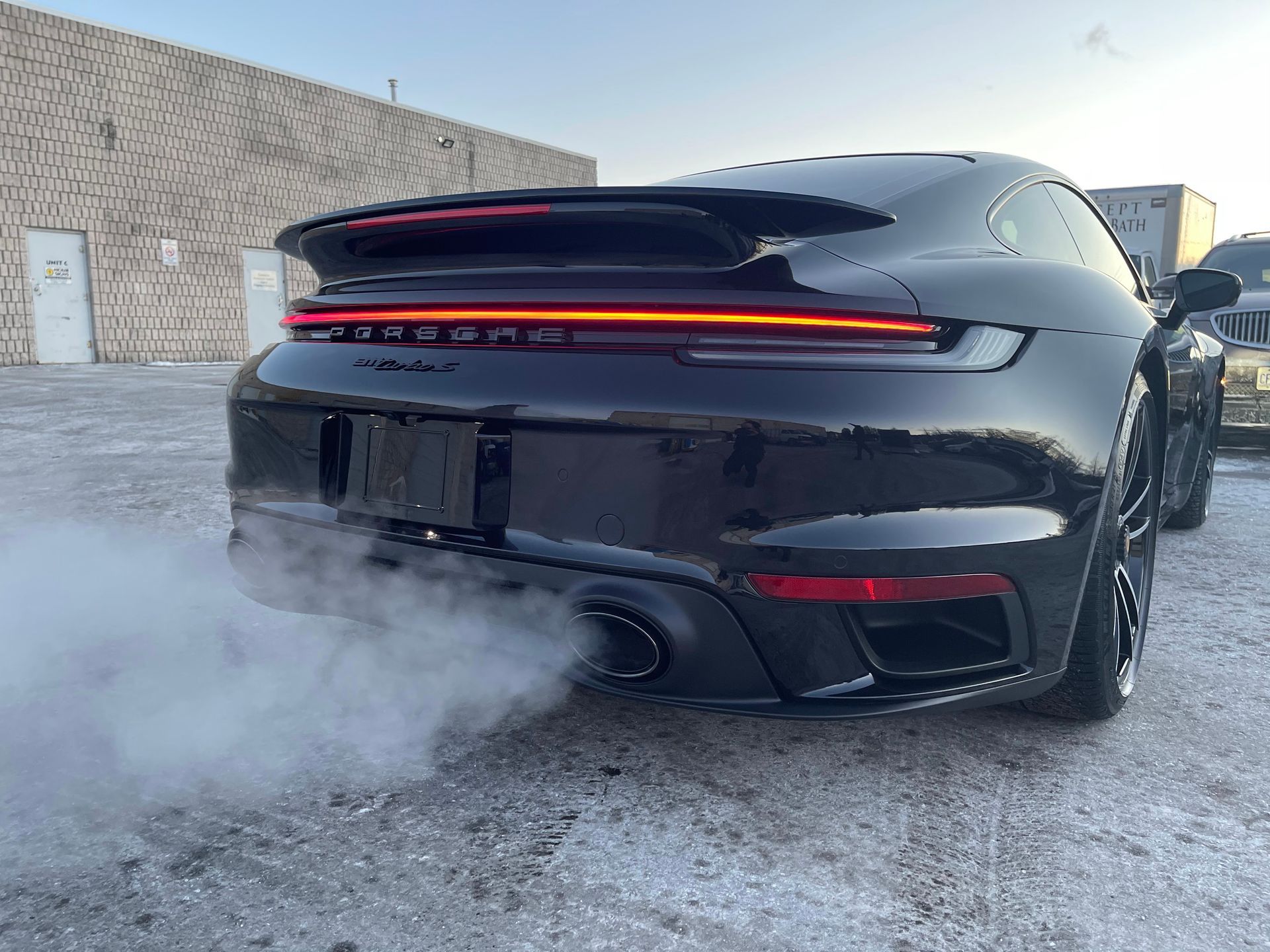 A black Porsche 911 parked outdoors with thick white exhaust smoke billowing from its tailpipe.