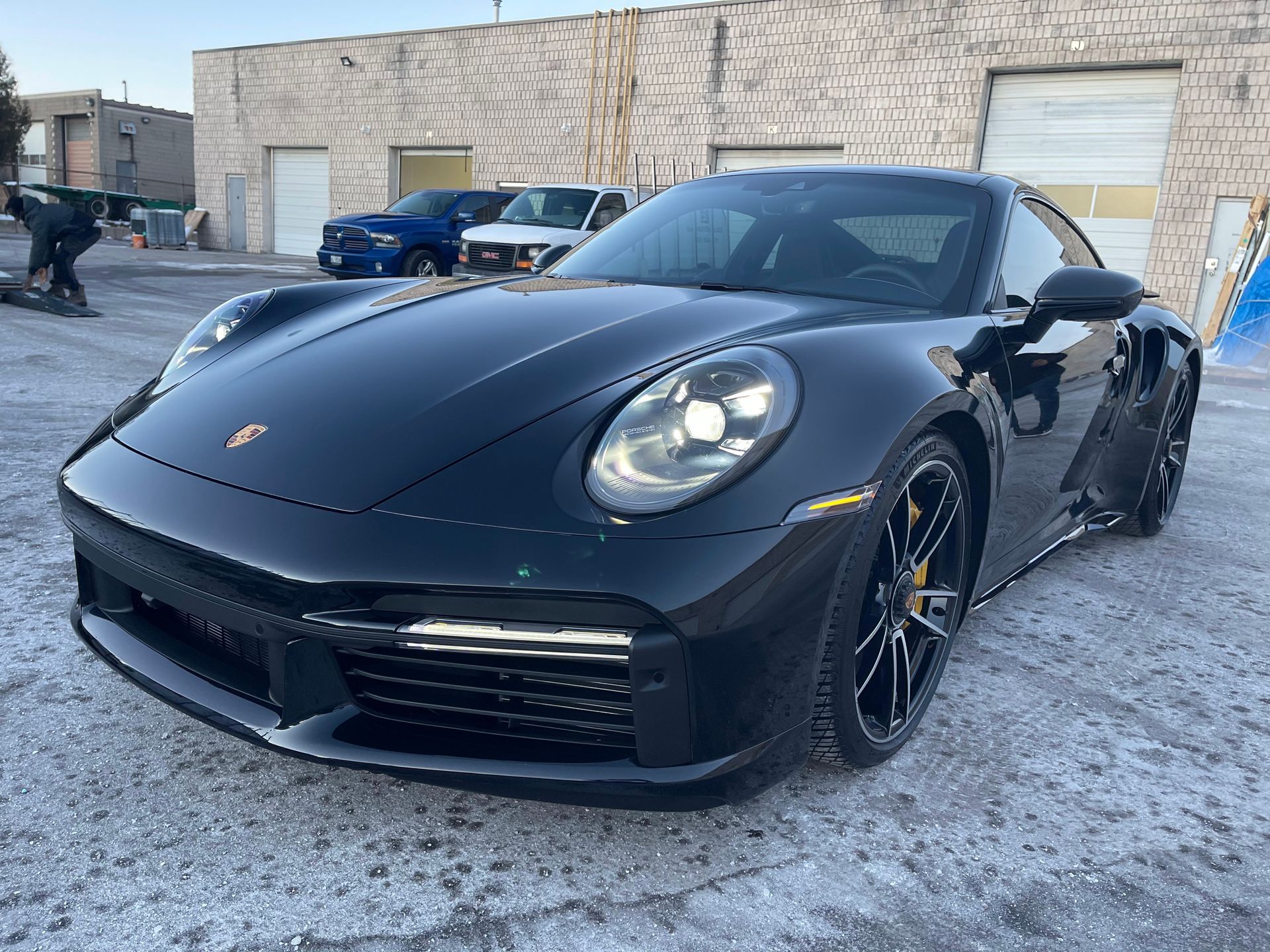 A black Porsche 911 parked on an asphalt lot in front of a tan brick industrial building.