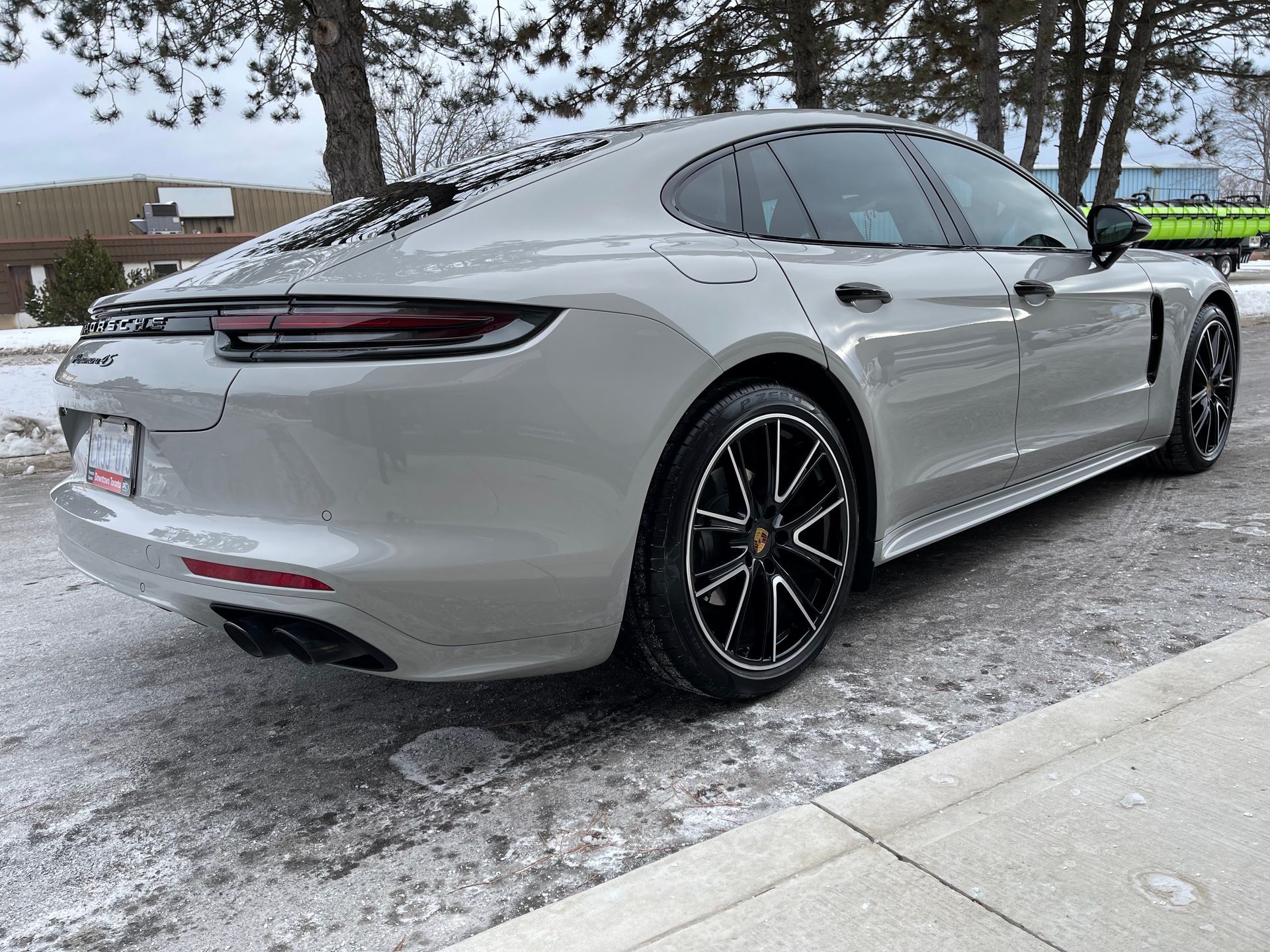 A grey Porsche Panamera sedan parked on a snowy, paved surface beside a curb, viewed from a rear three-quarter angle.