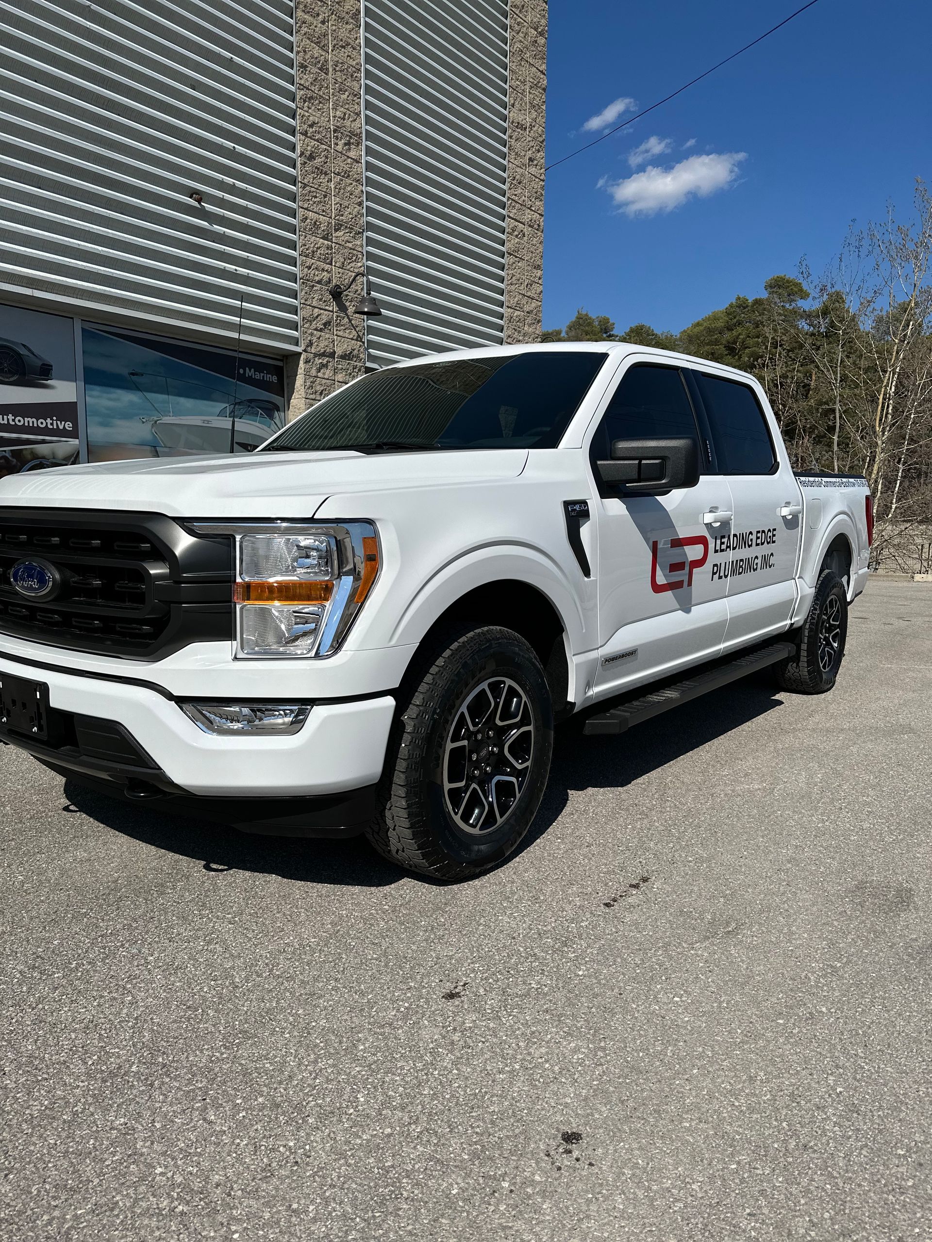 A white Ford F-150 pickup truck with black trim and company branding parked on a gravel lot under a clear blue sky.