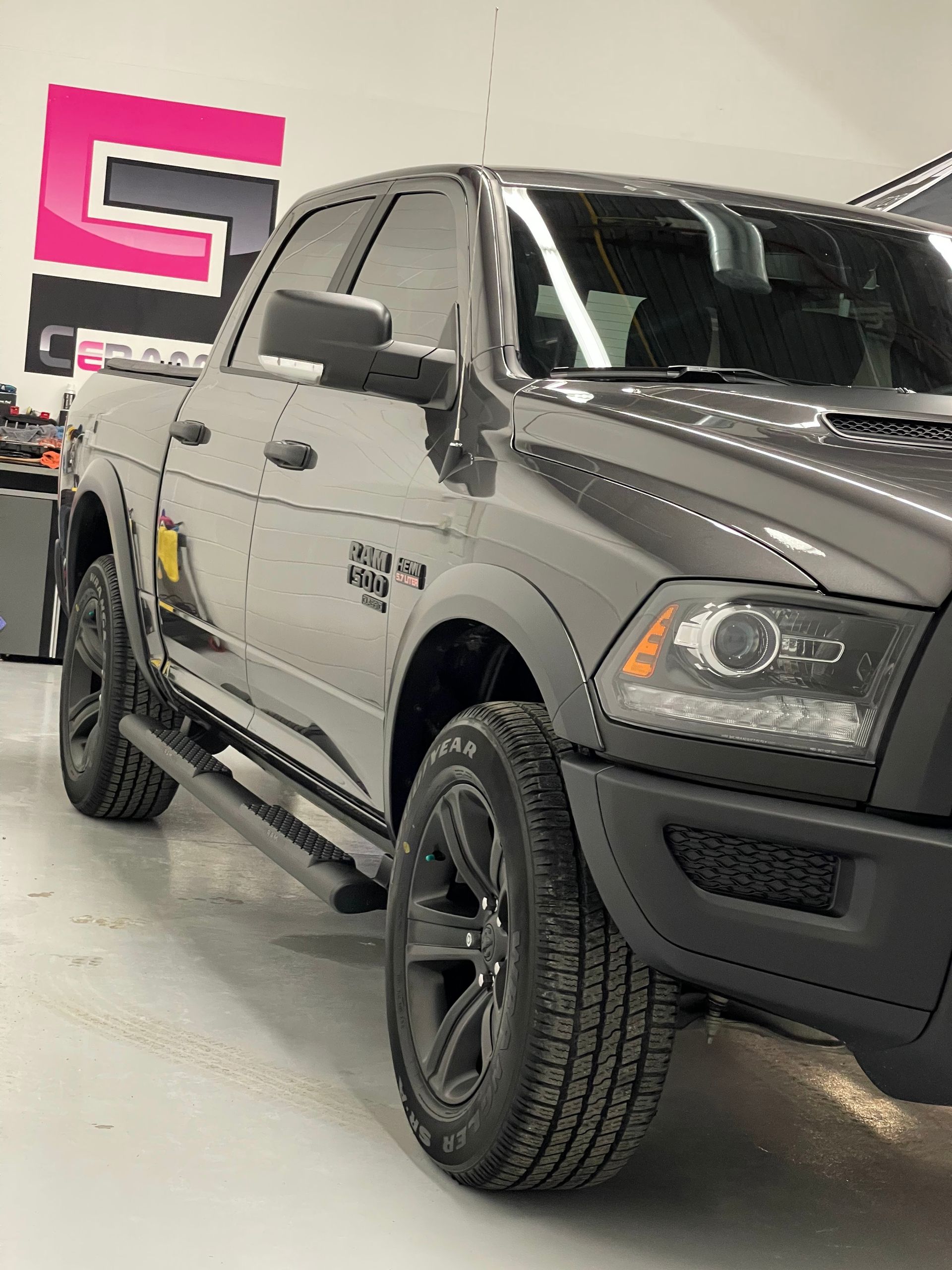 A dark gray pickup truck parked in a well-lit workshop with a prominent pink and black logo on the wall behind it.