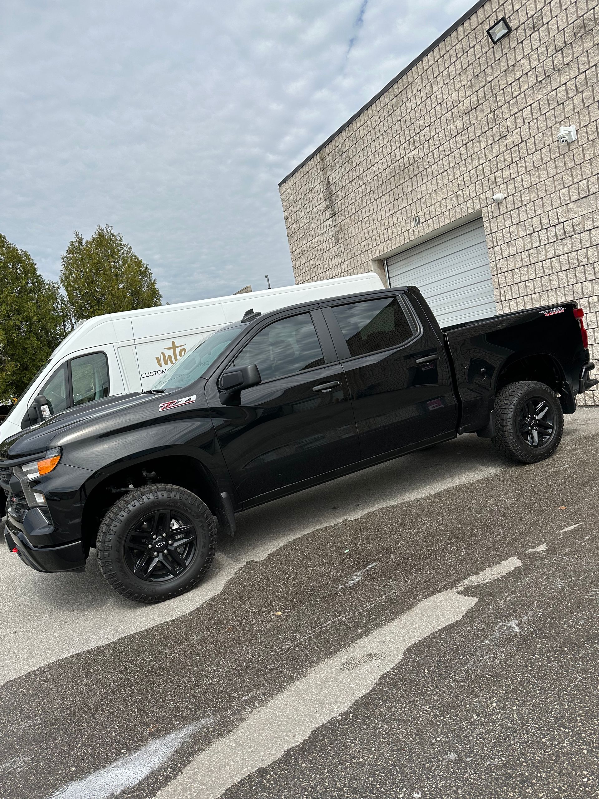 A black Chevrolet Silverado pickup truck parked outside a building with a white van in the background.