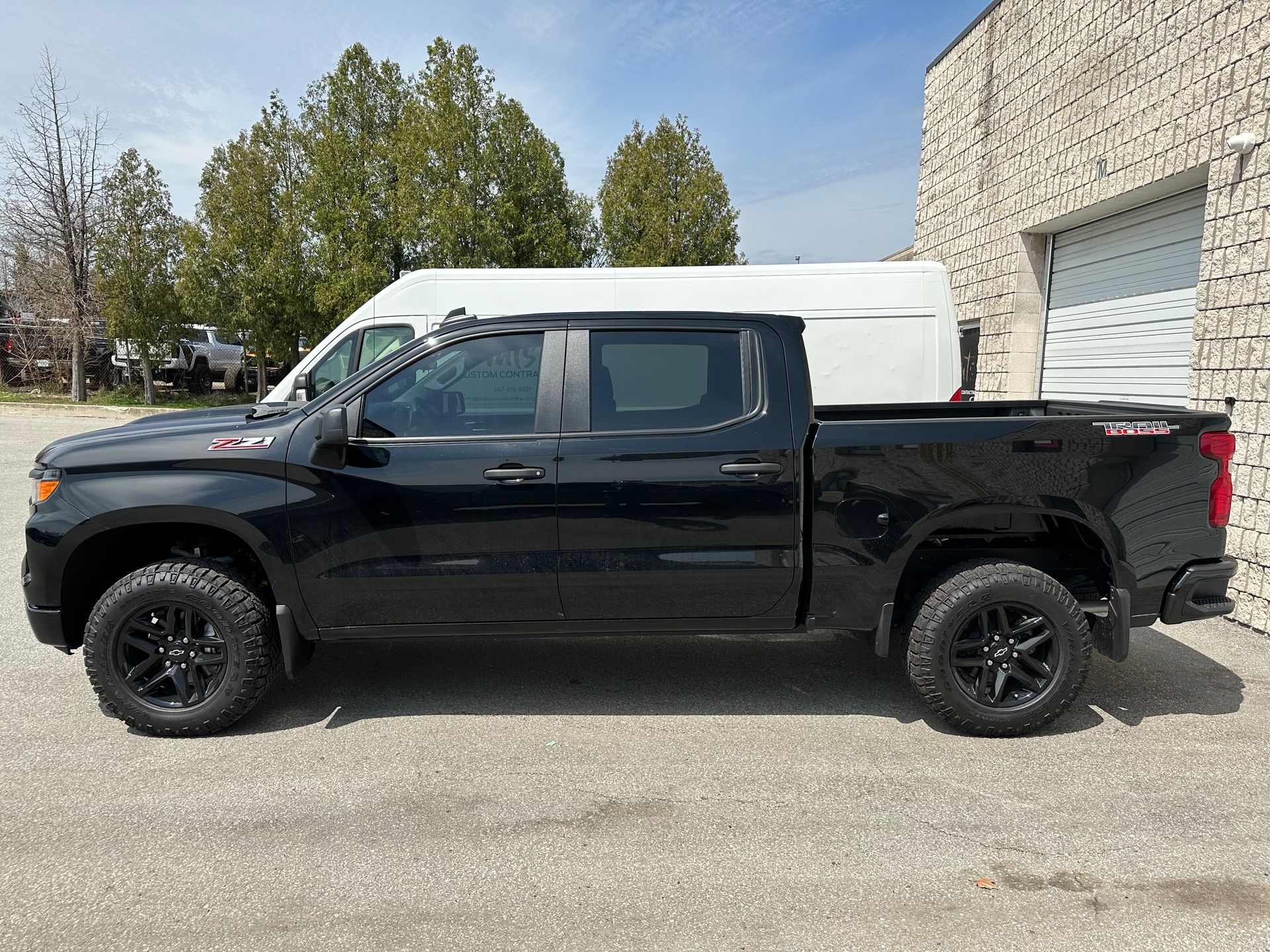 A black crew cab pickup truck parked outdoors in front of a white van and a stone building.