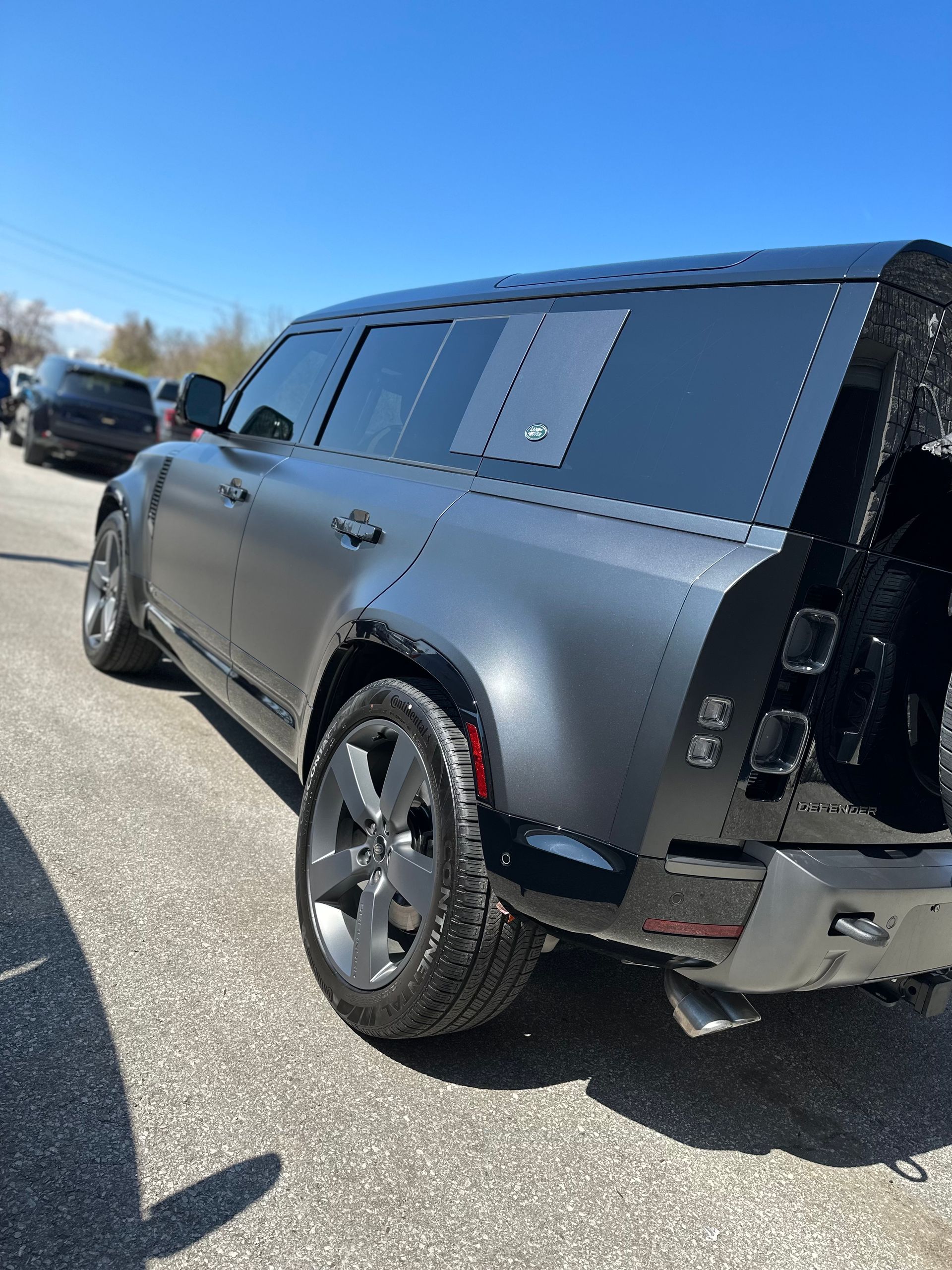 A matte black Land Rover Defender parked outdoors on a sunny day, viewed from the rear passenger side.