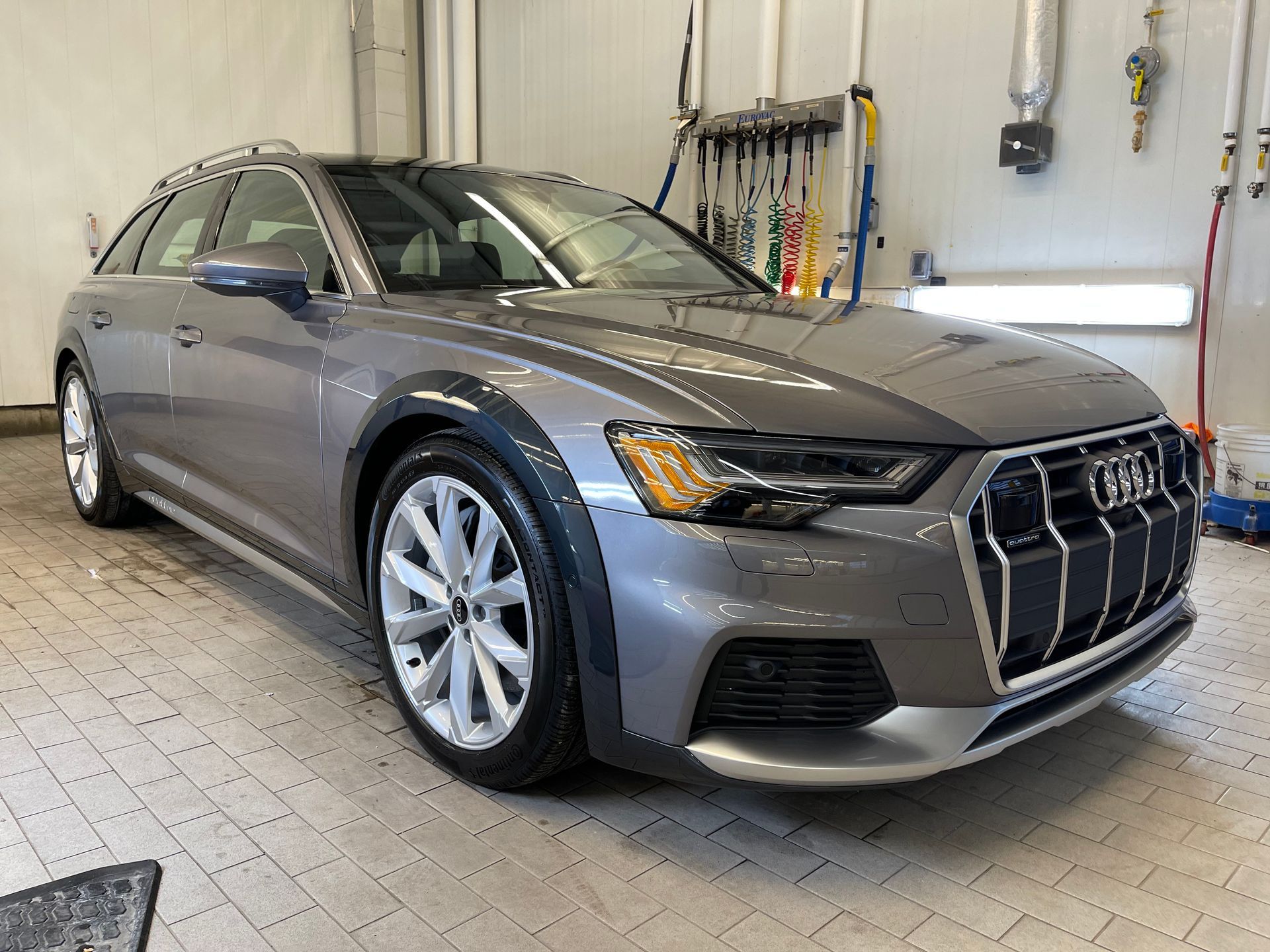 A grey Audi A6 Allroad wagon parked inside a brightly lit, tiled service garage.