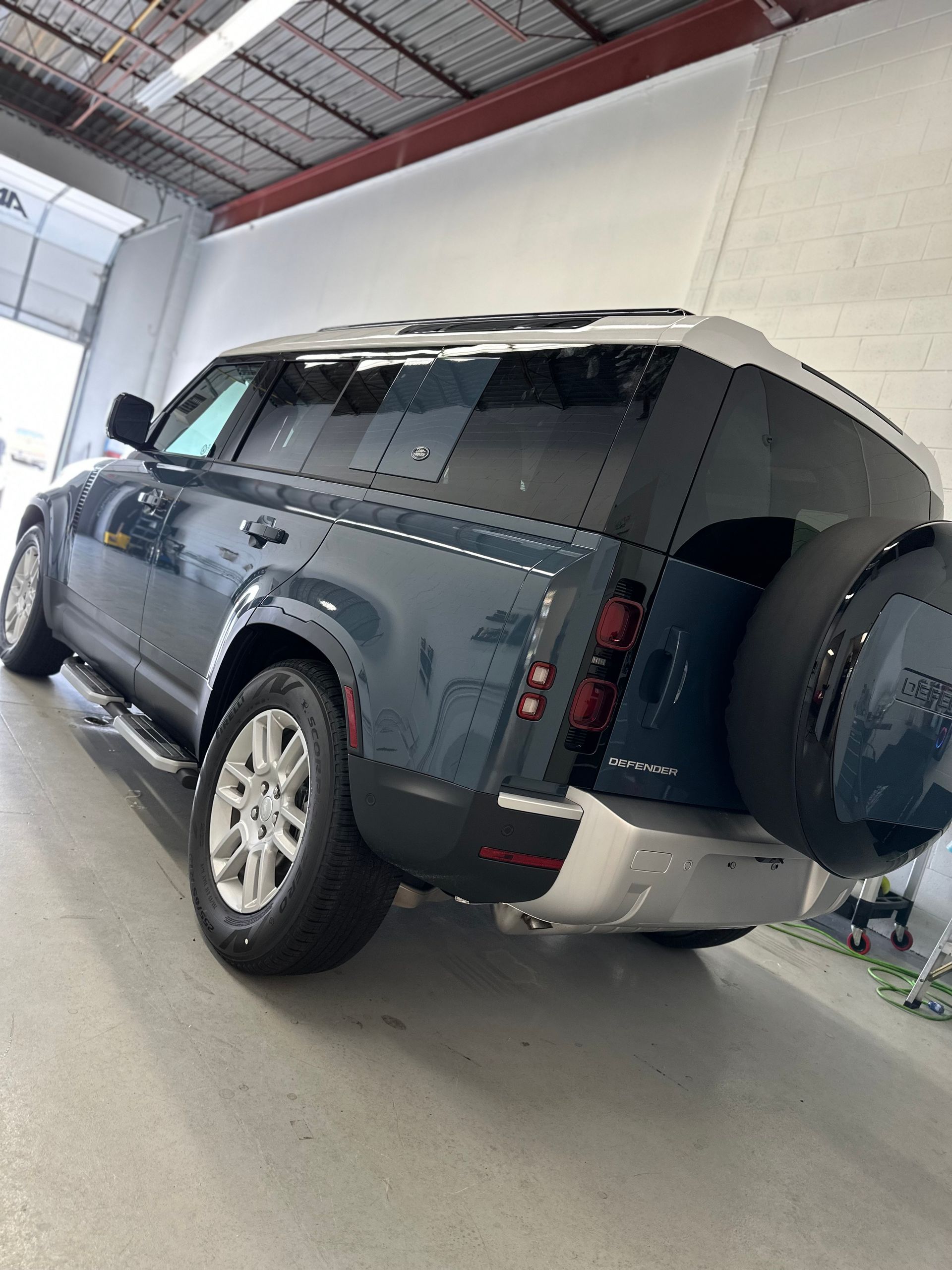 A blue Land Rover Defender parked inside a well-lit garage, viewed from the rear three-quarter angle.