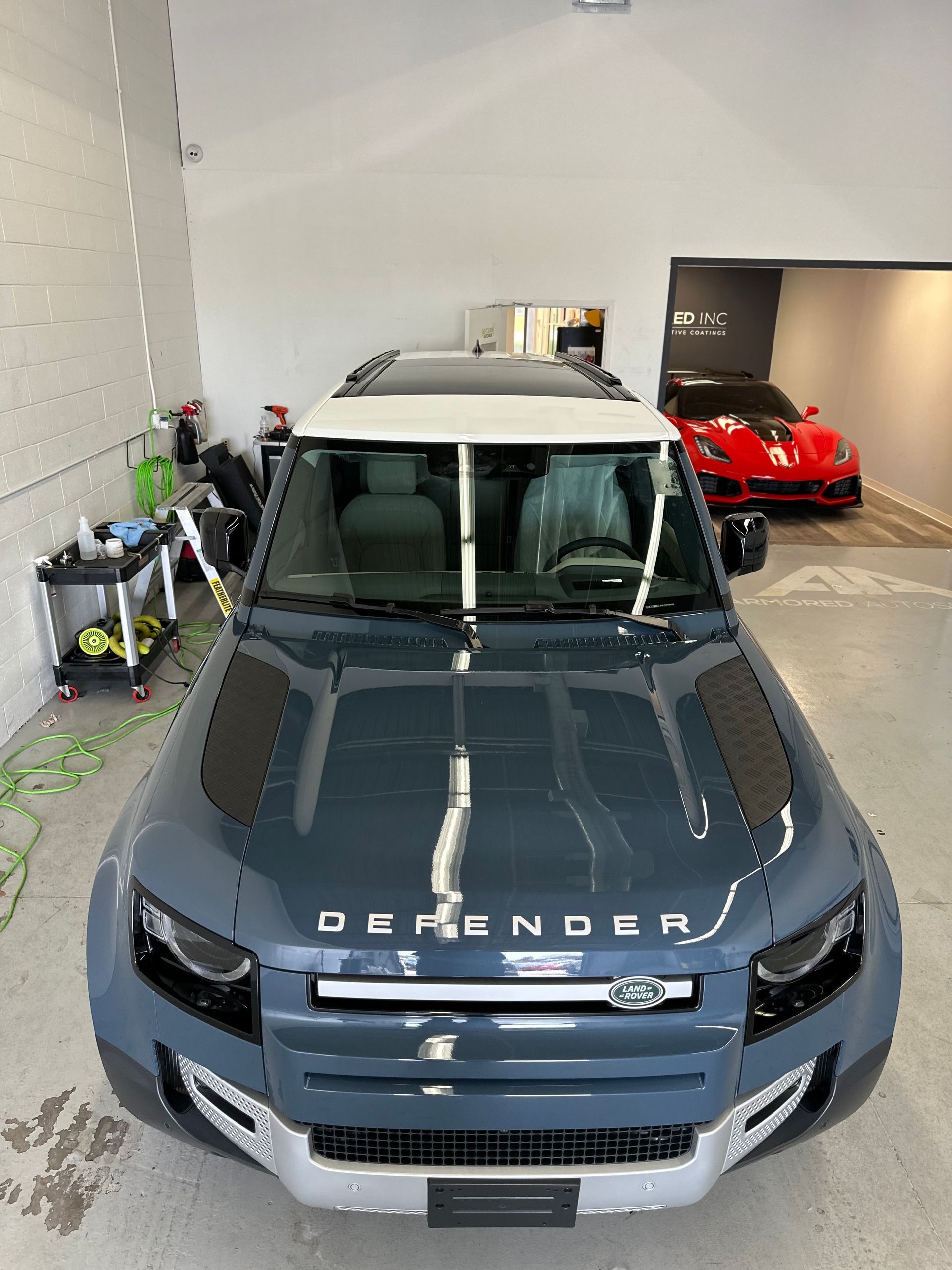 A gray Land Rover Defender with a white roof in a garage, with a blurred red sports car visible in the background.