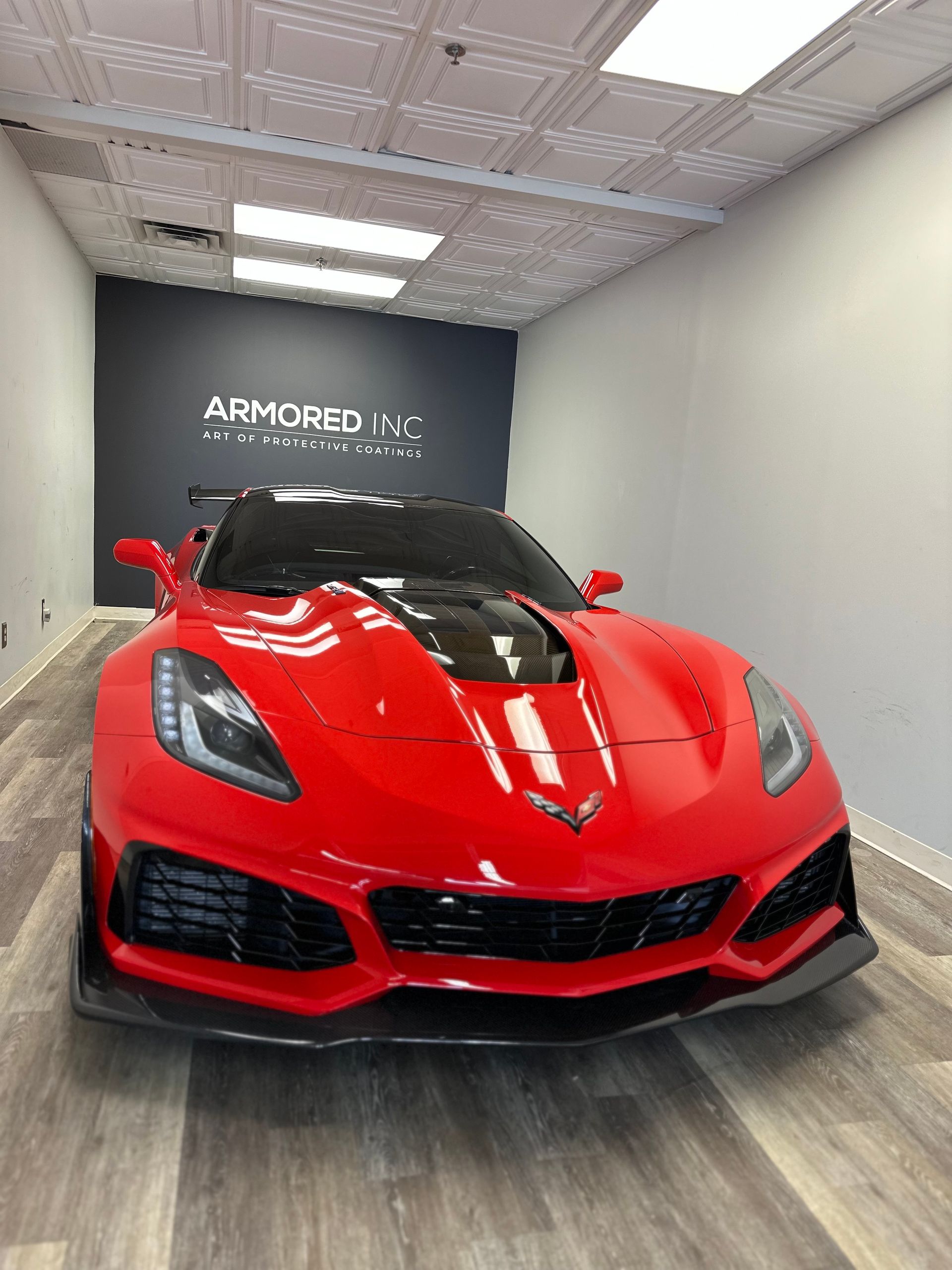 Bright red Chevrolet Corvette ZR1 with a black hood parked in a showroom with light wood floors and a dark feature wall.