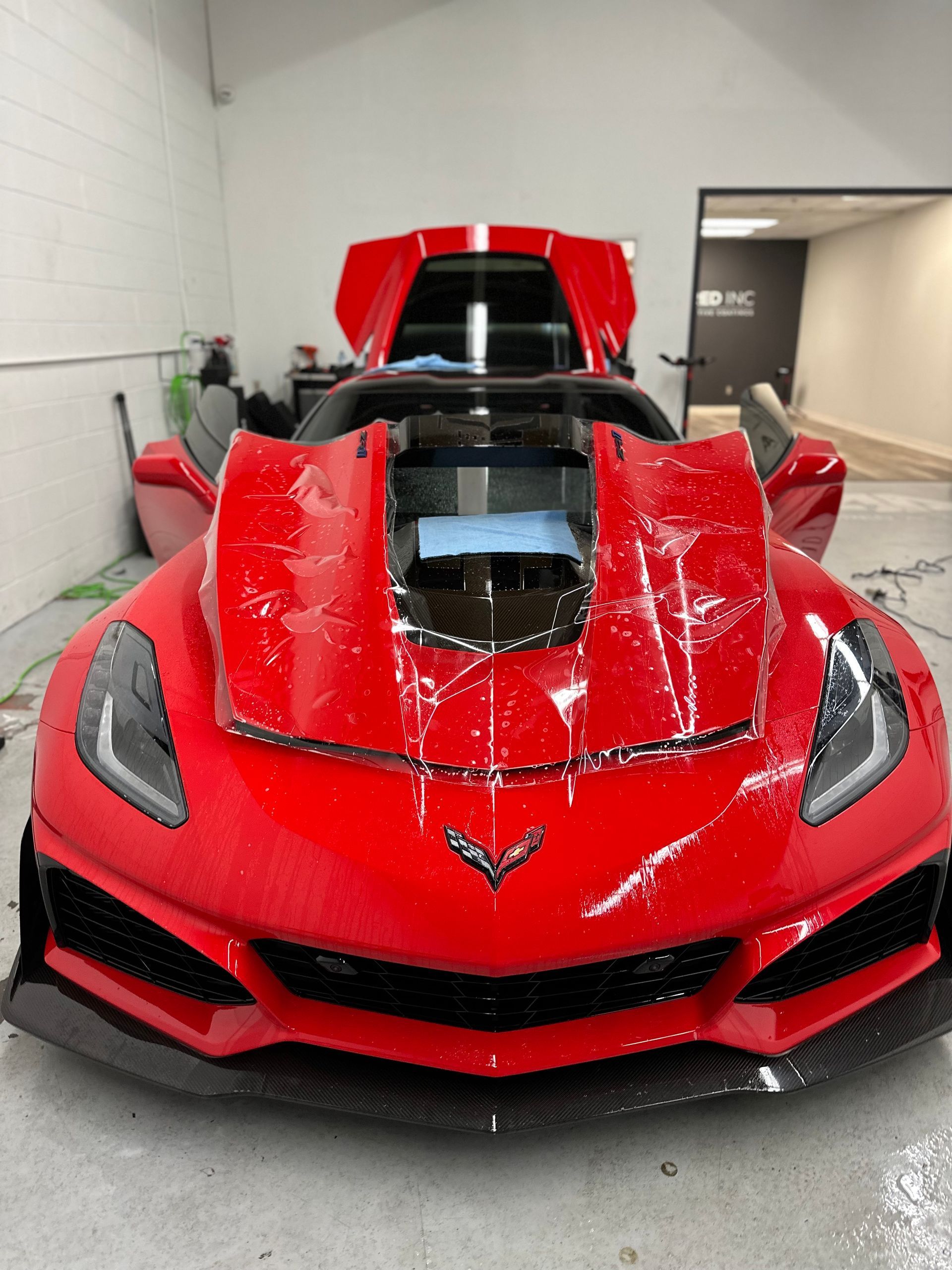 Bright red Corvette parked in a garage with its hood and doors open, undergoing automotive detailing.