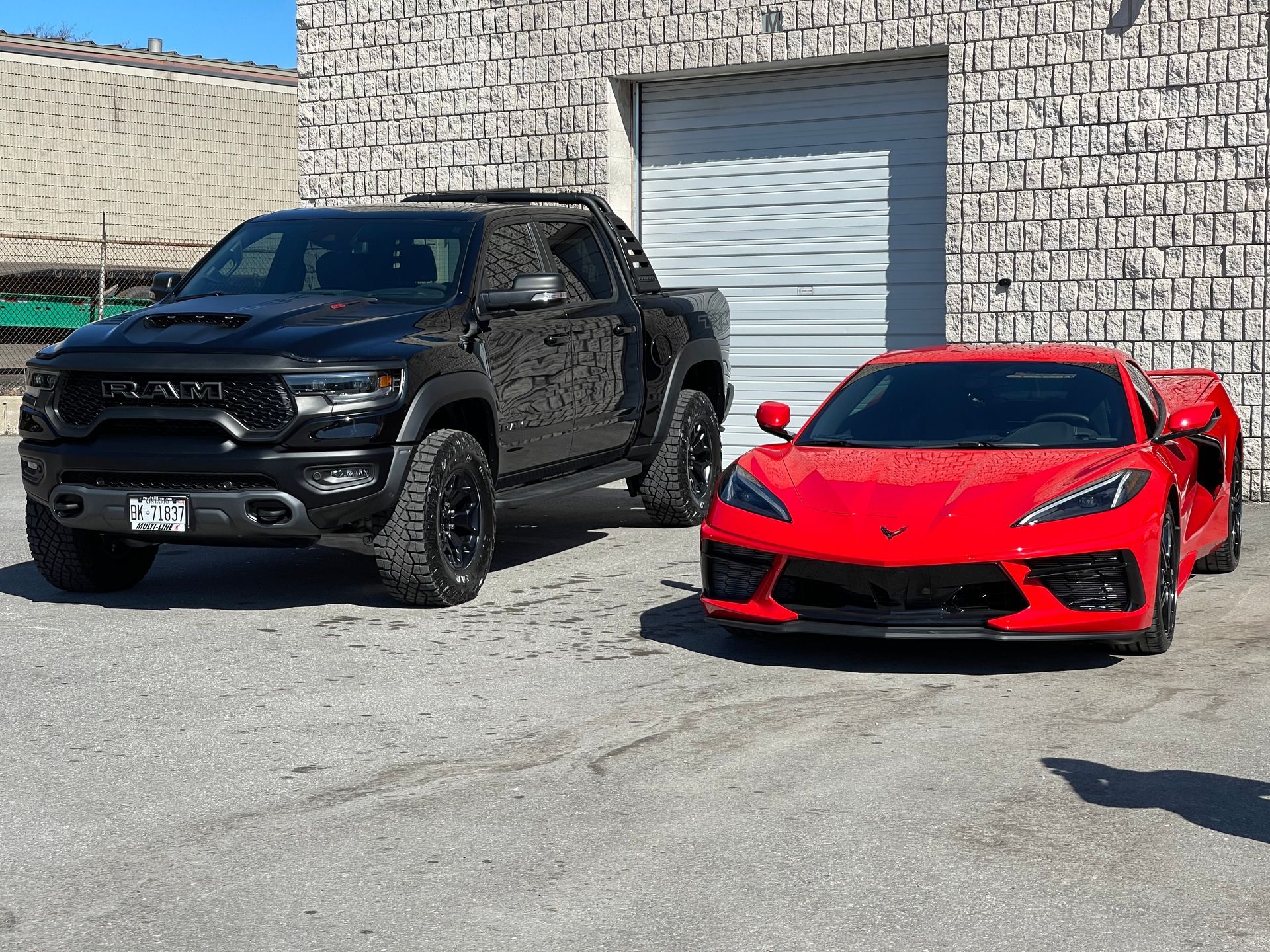 A black RAM TRX truck and a bright red Chevrolet Corvette parked side-by-side on an asphalt lot in front of a building.