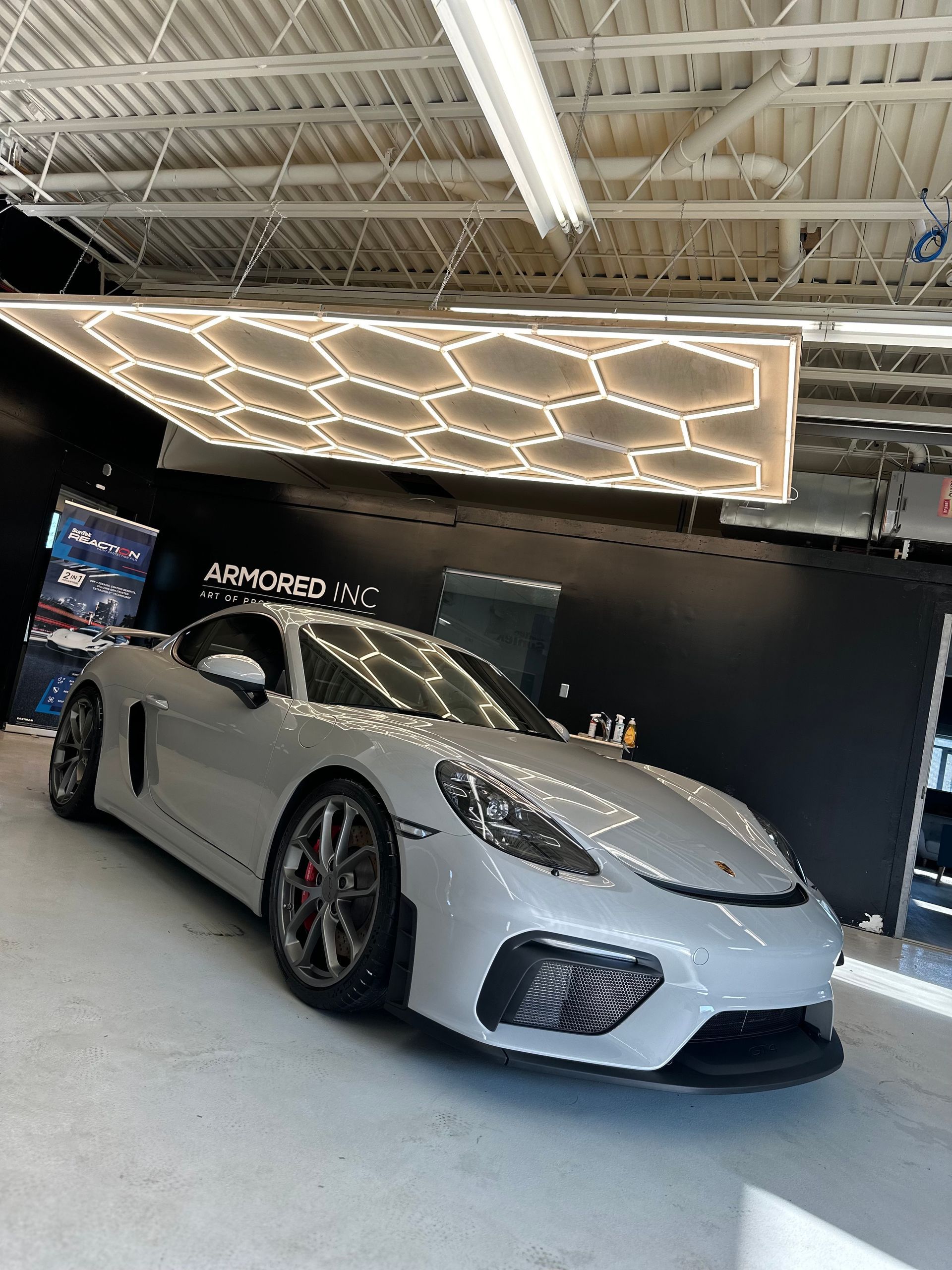 A grey Porsche sports car parked in an indoor garage under a large, illuminated hexagonal ceiling light fixture.