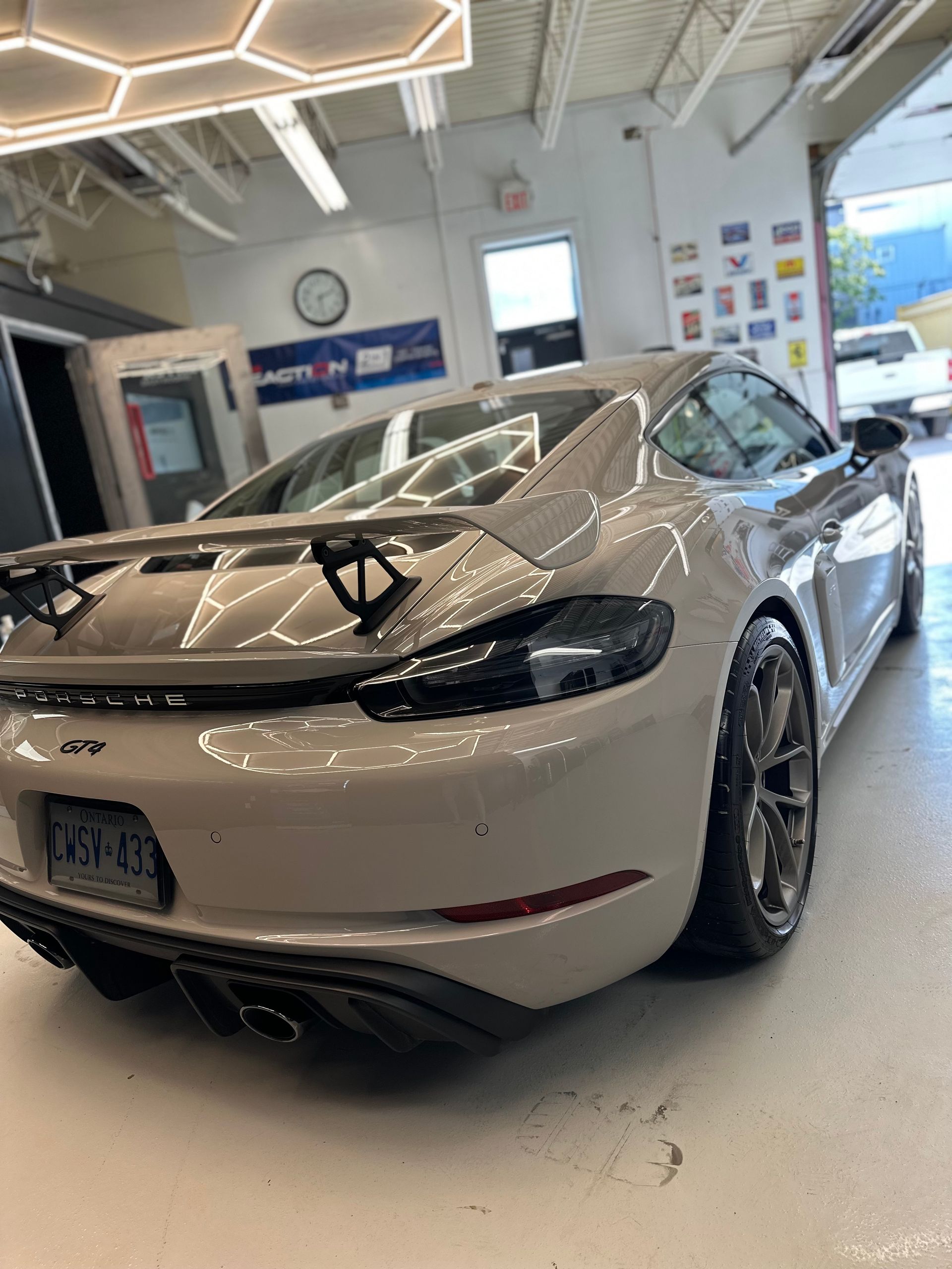 Chalk-gray Porsche 718 Cayman GT4 with a rear wing and black diffuser parked inside a brightly lit garage.