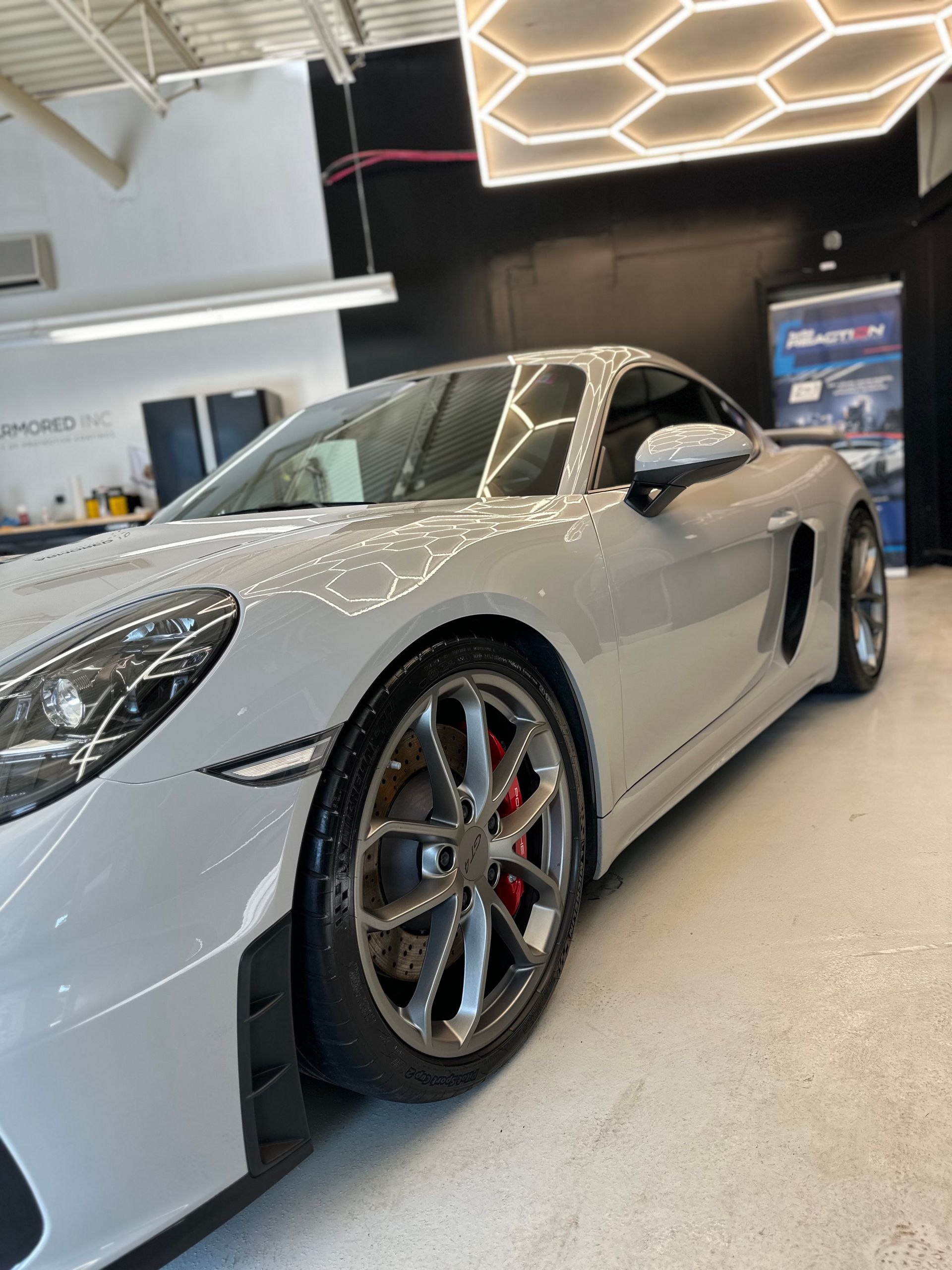 A light grey Porsche sports car is parked inside a brightly lit automotive studio with hexagonal ceiling lights.