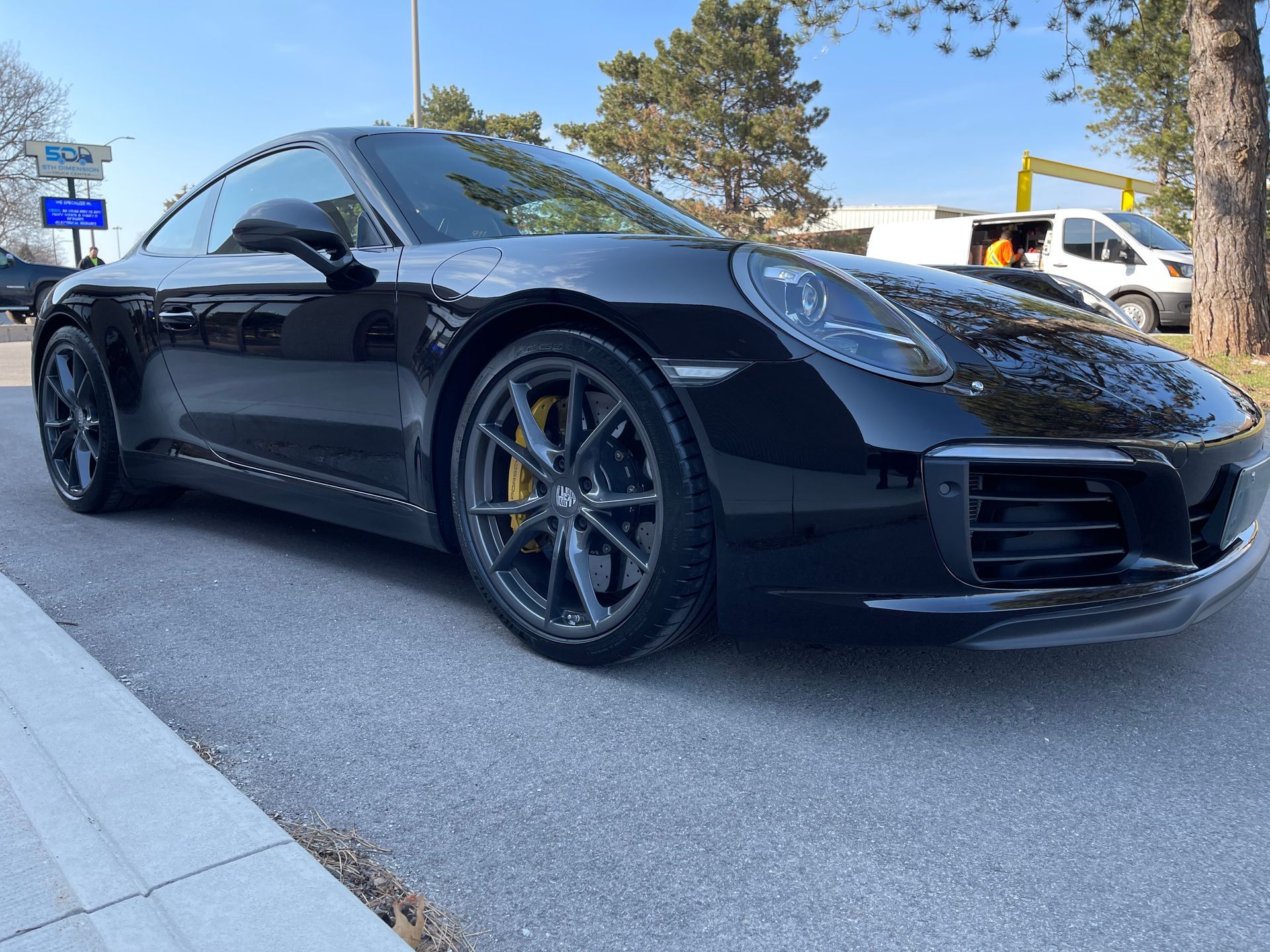 A black Porsche 911 parked on an asphalt street with a white van and trees in the background.