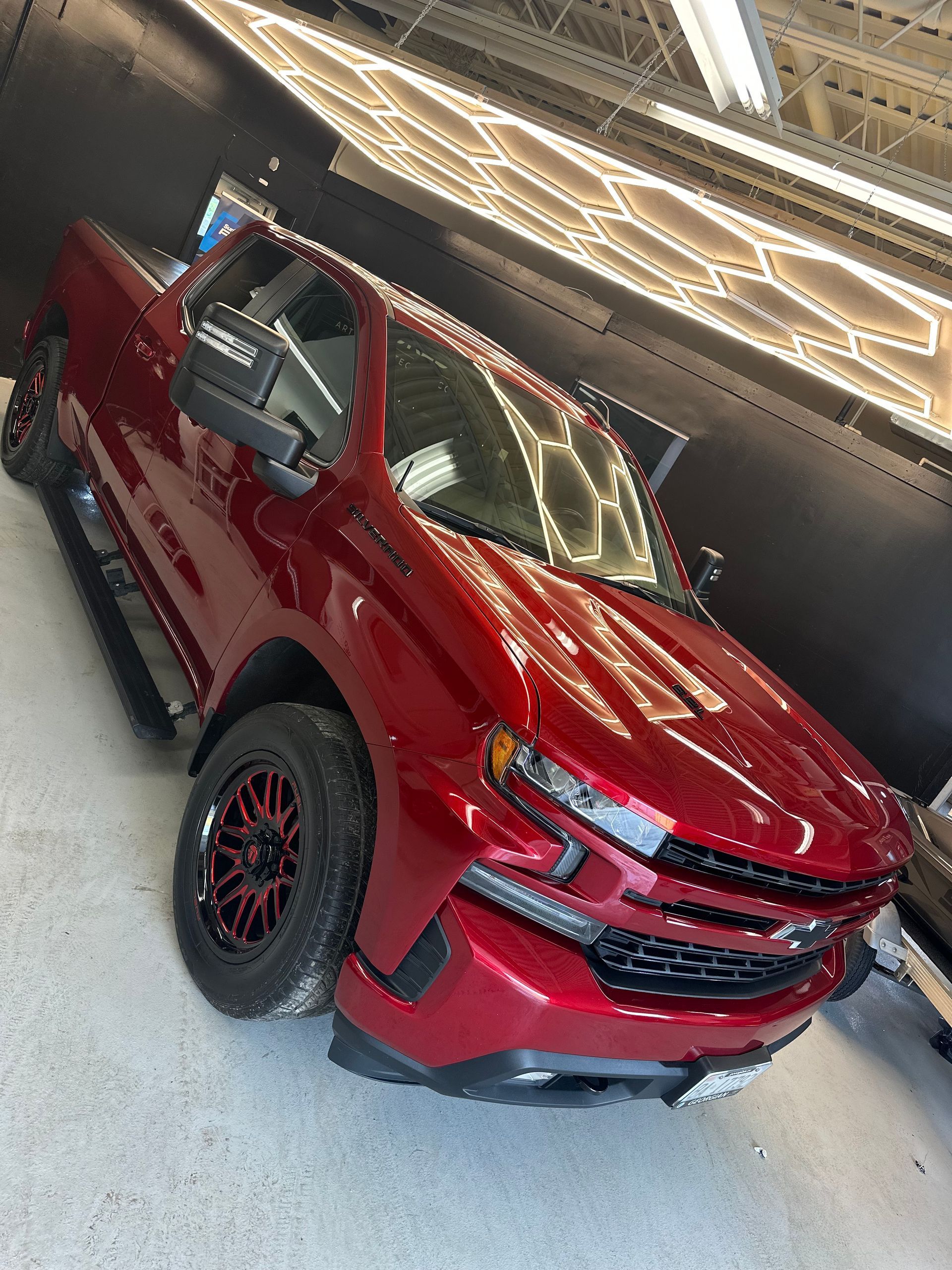 A glossy cherry red Chevrolet pickup truck parked in a well-lit indoor garage with a honeycomb ceiling pattern.