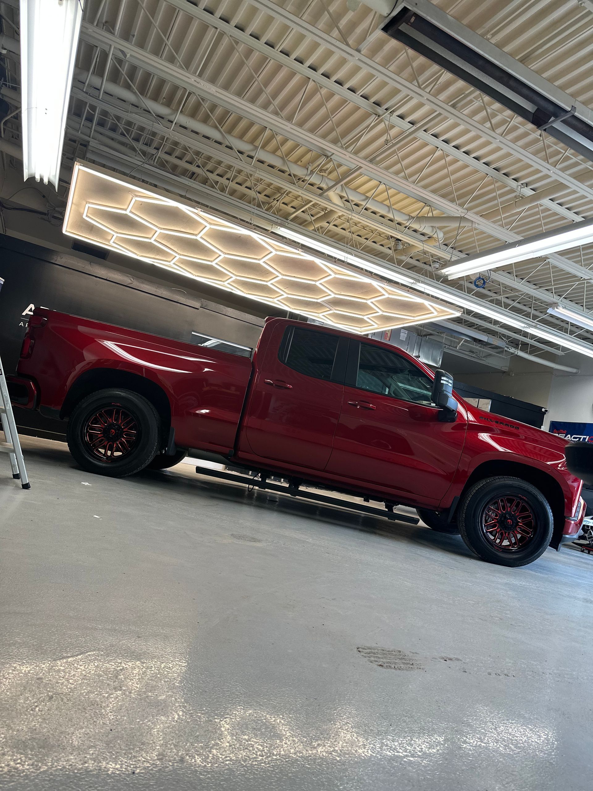 A side profile of a red pickup truck parked in a workshop with overhead honeycomb-patterned LED lighting.
