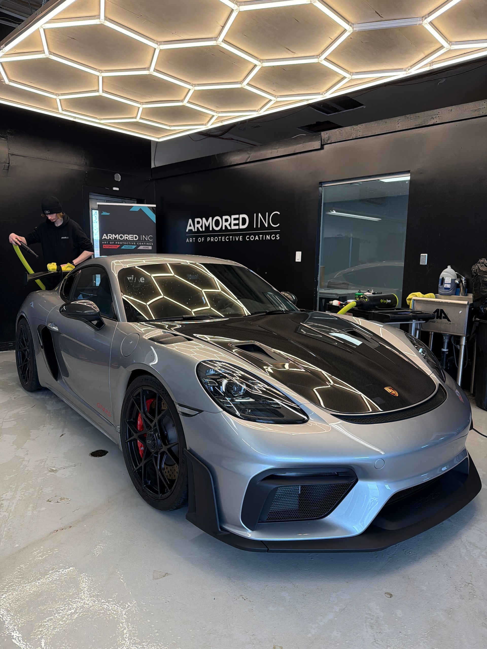 A silver Porsche sports car with a carbon fiber hood parked in an auto shop garage with honeycomb ceiling lights.