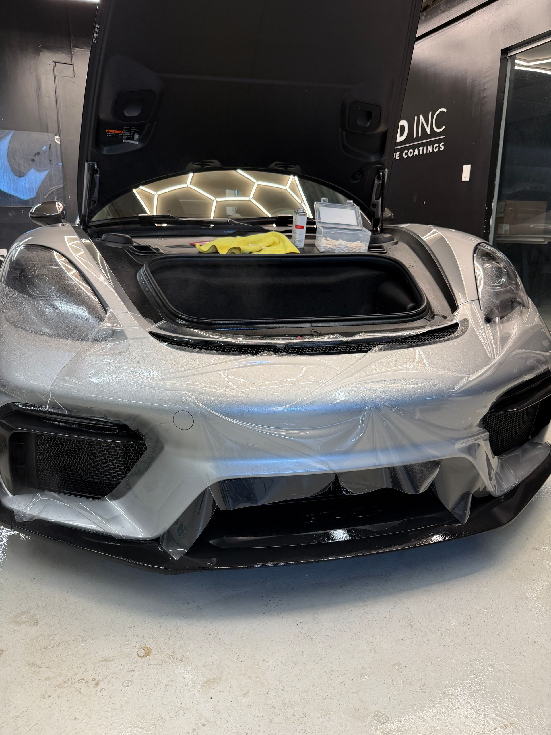 A silver Porsche sports car inside a workshop with its hood open and paint protection film being applied.