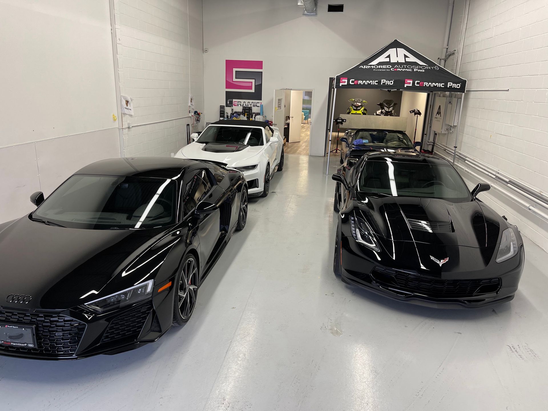 Three sports cars, including two black ones and a white one, parked inside a brightly lit professional auto shop.