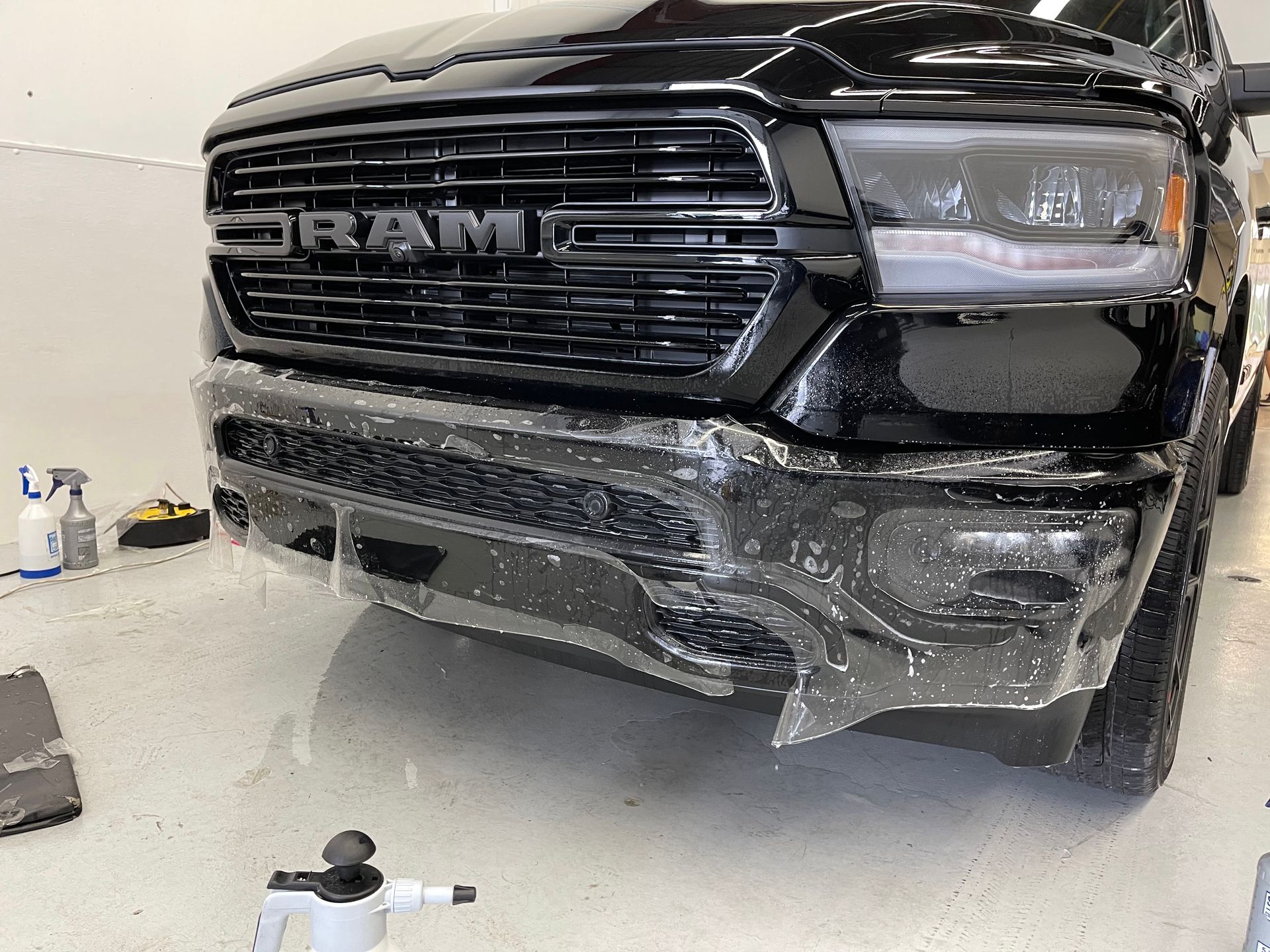 A black RAM truck bumper partially covered with a clear paint protection film during an installation in a shop.