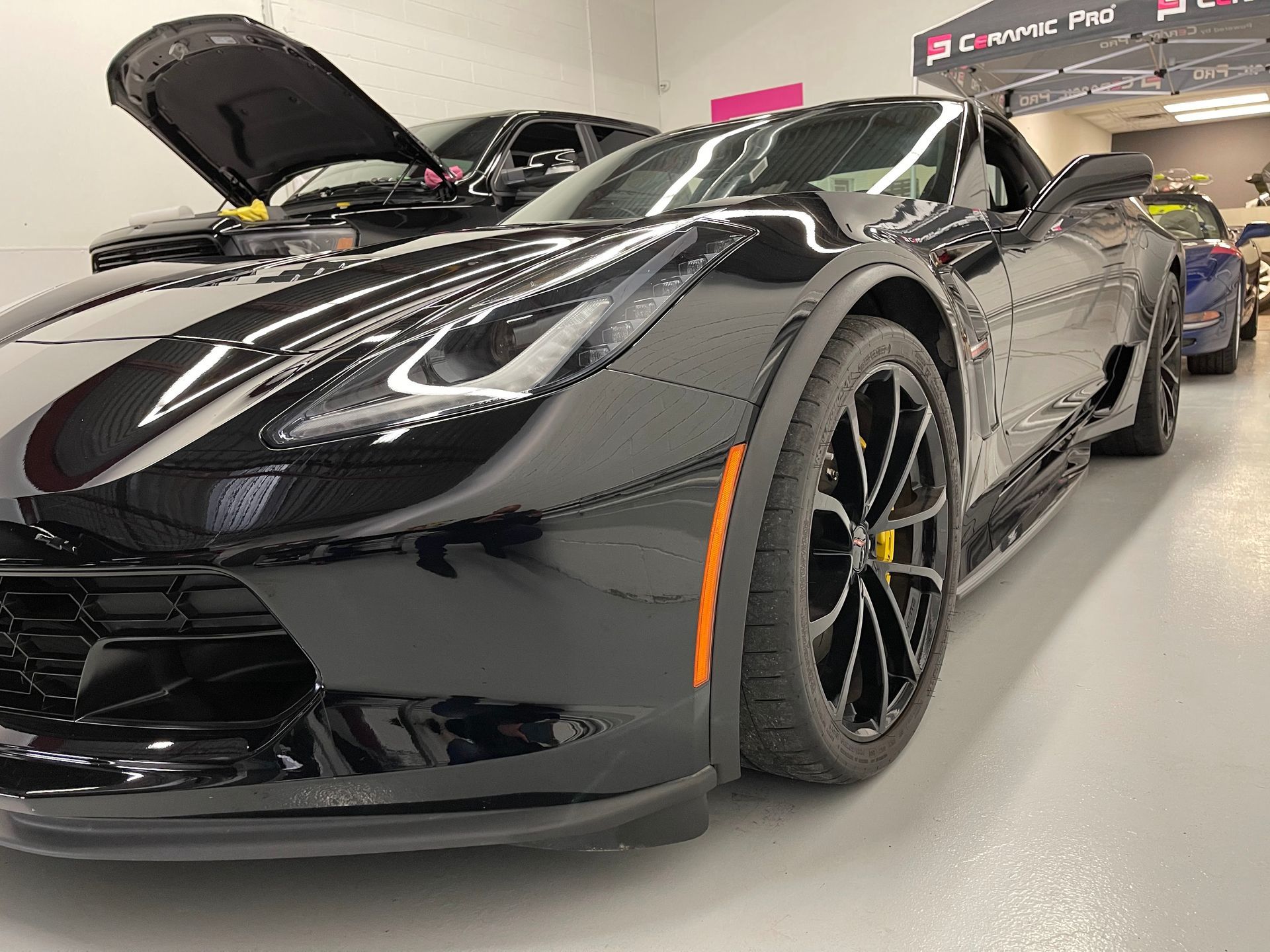 A close-up, low-angle shot of a black Chevrolet Corvette parked inside a showroom with another car in the background.