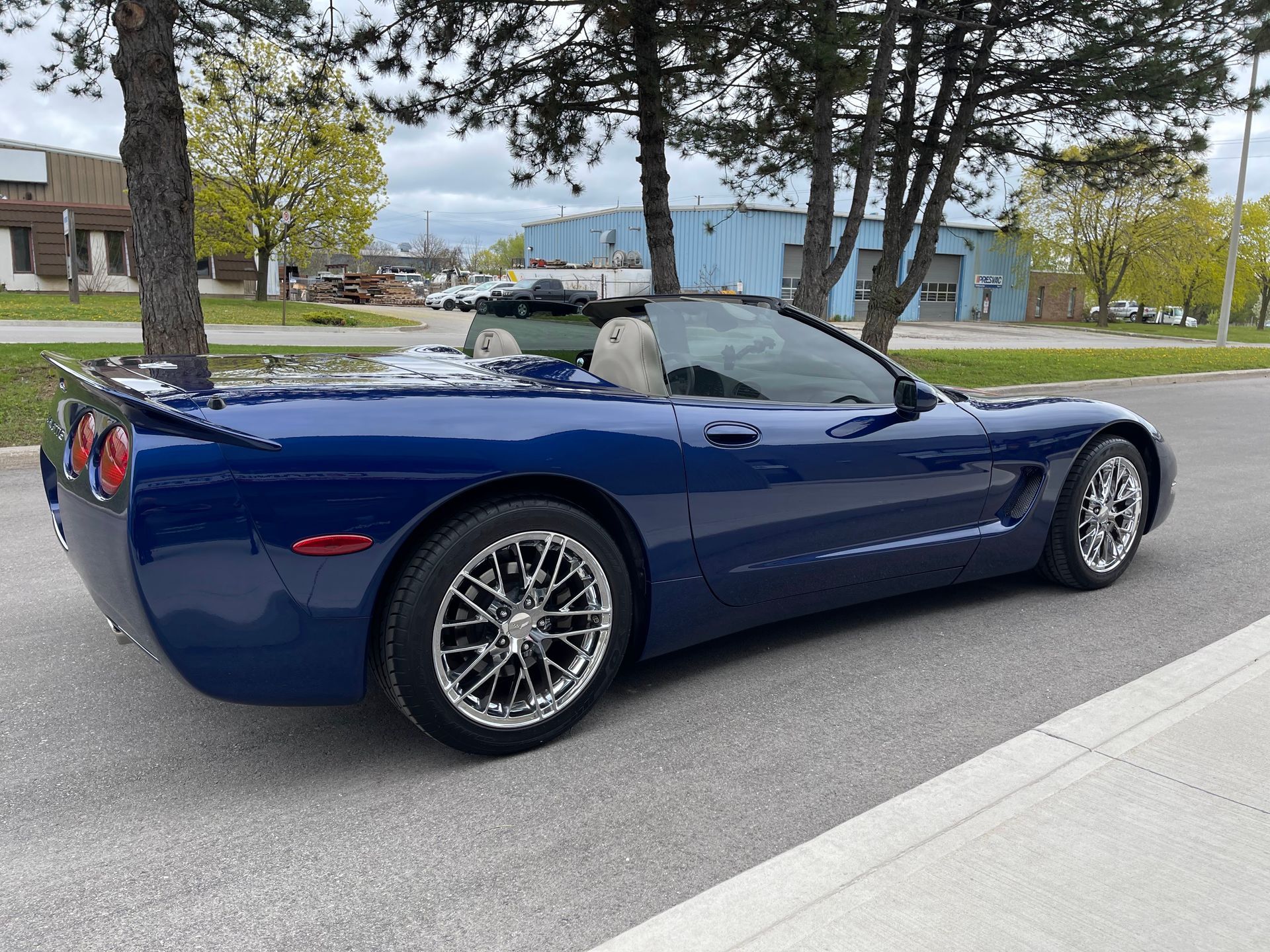 A dark blue convertible Chevrolet Corvette parked on an asphalt street with trees and a light blue building in the back.