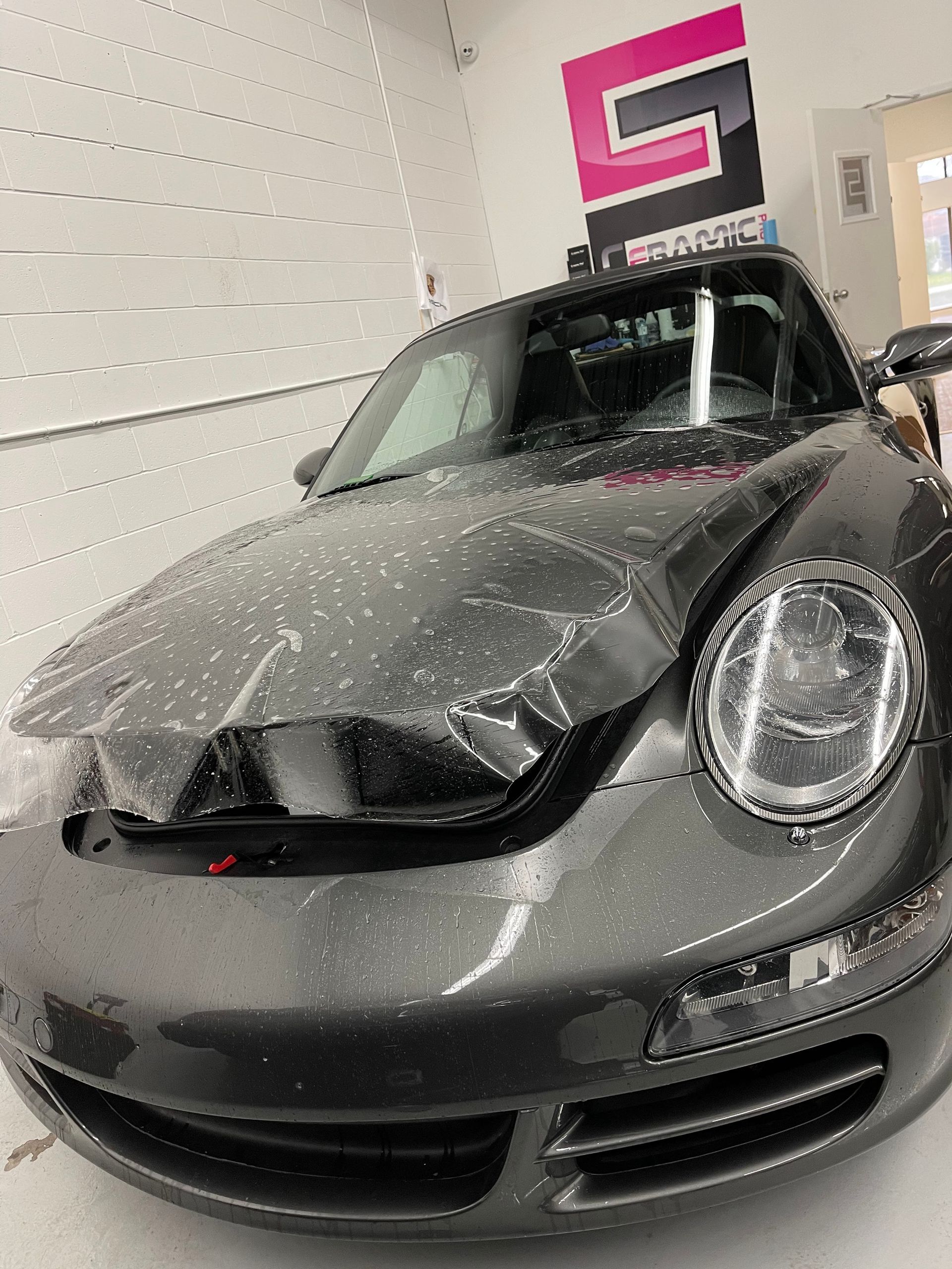 A dark gray Porsche sports car undergoing a paint protection film installation on its hood in a workshop.